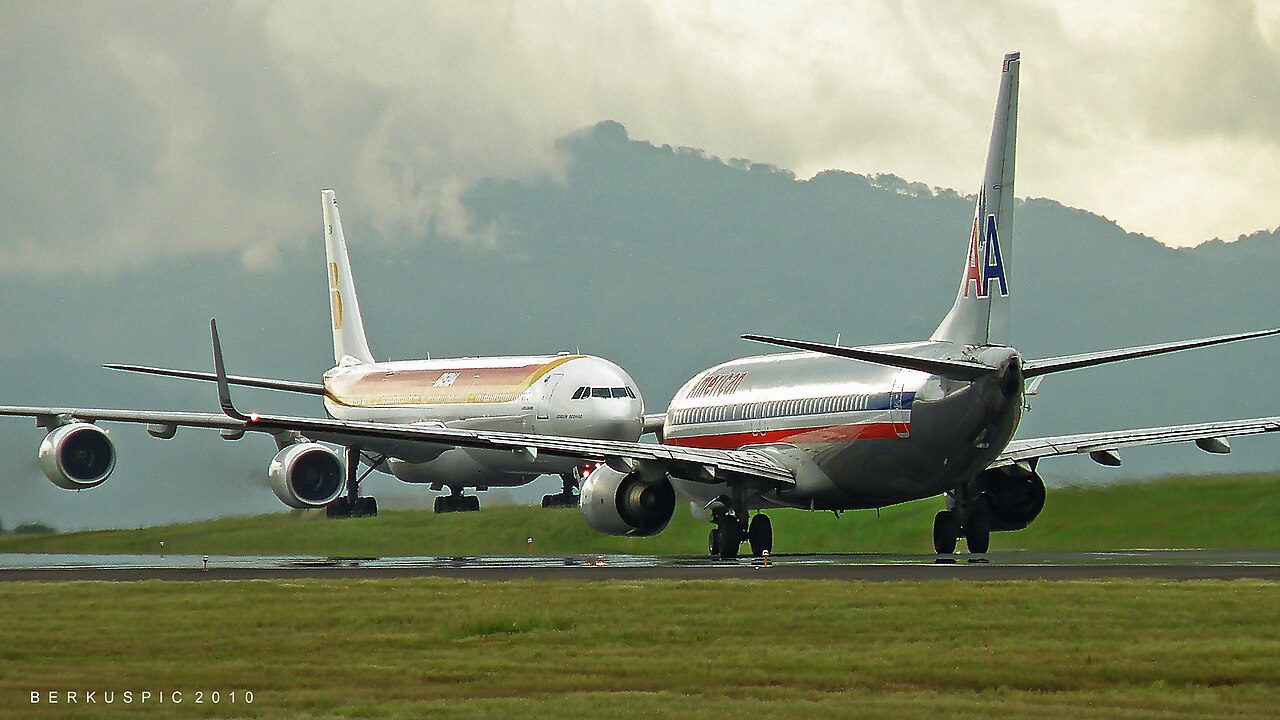 An American Airlines Boeing 737 and an Iberia Airbus A340 on the runway at Juan Santamaría International Airport, with green hills under cloud behind.