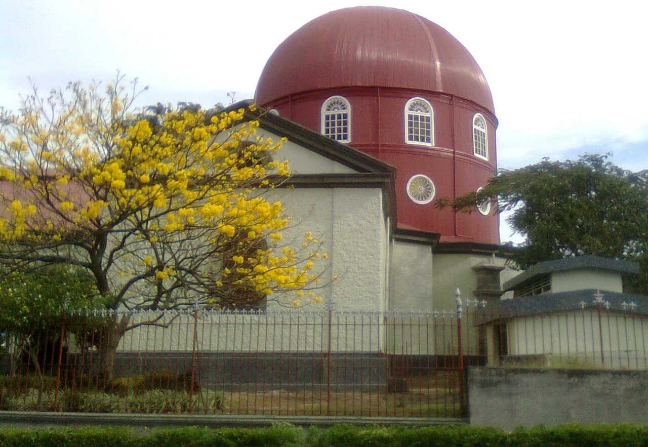 The red metal dome of the cathedral in Alajuela, Costa Rica, behind a white church wall, an iron fence and a tree with yellow blossoms.
