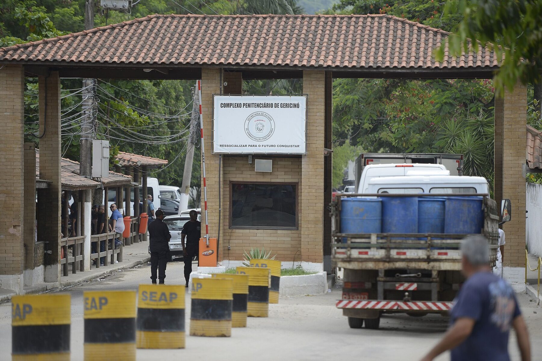 Entrance gate of the Gericinó prison complex in Rio de Janeiro, with guards, a truck and yellow SEAP bollards