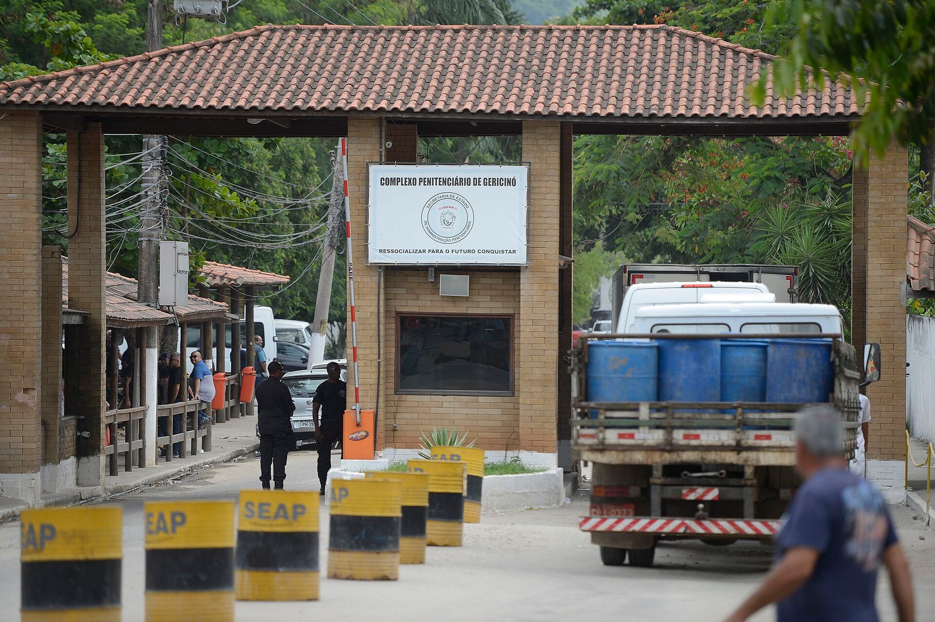 Entrance gate of the Gericinó prison complex in Rio de Janeiro, with guards, a truck and yellow SEAP bollards