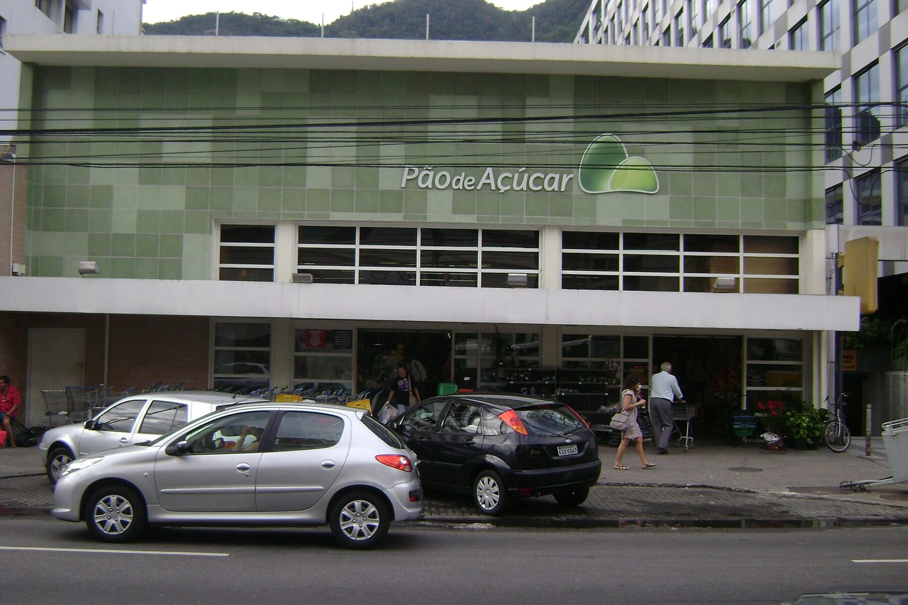 Front of a Pão de Açúcar supermarket with a green tiled facade, cars parked outside and shoppers on the pavement