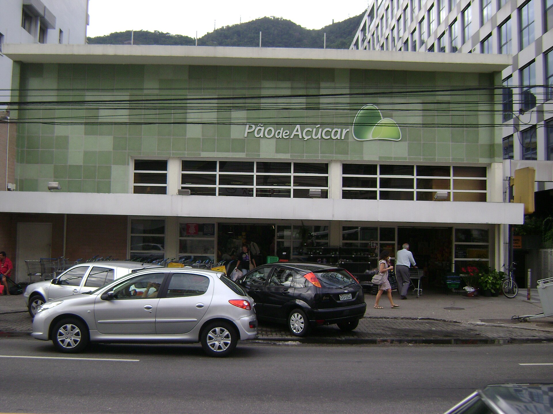 Front of a Pão de Açúcar supermarket with a green tiled facade, cars parked outside and shoppers on the pavement