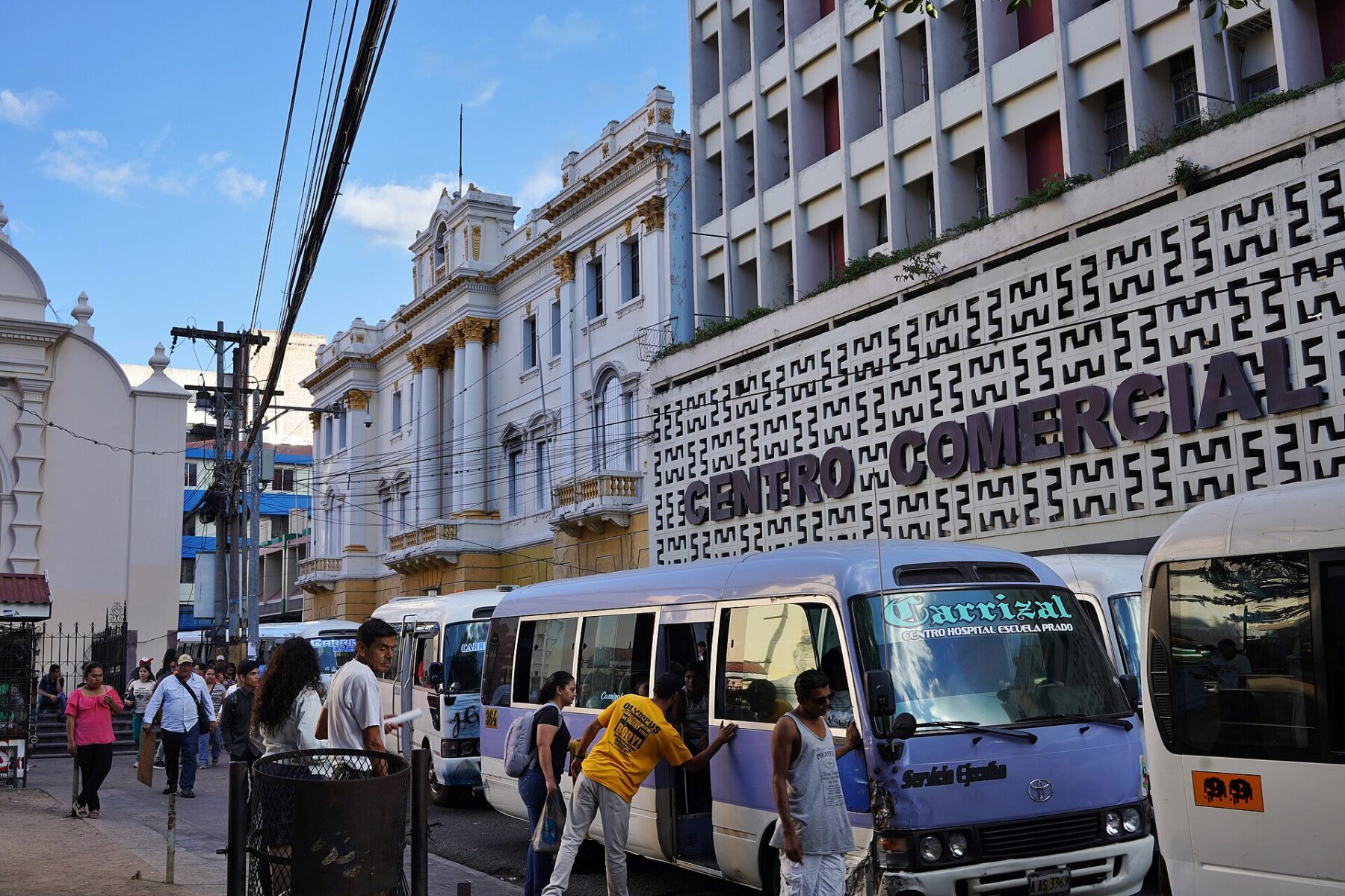 Minibuses and pedestrians on a street in central Tegucigalpa, with a cream colonial building and a shopping-centre facade reading Centro Comercial