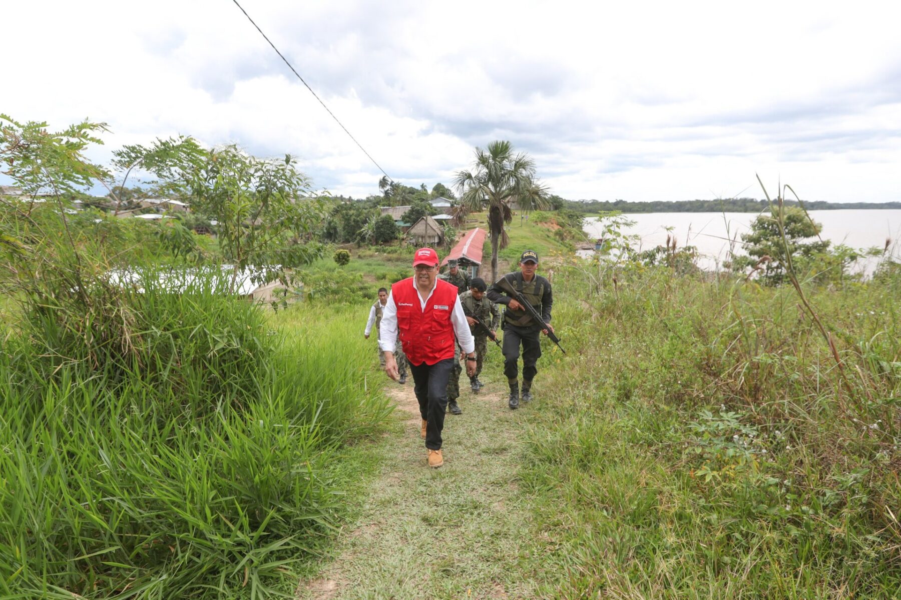 An official in a red vest and a red cap walks along a grassy riverside path, followed by armed soldiers in camouflage, with a village and a wide river behind them