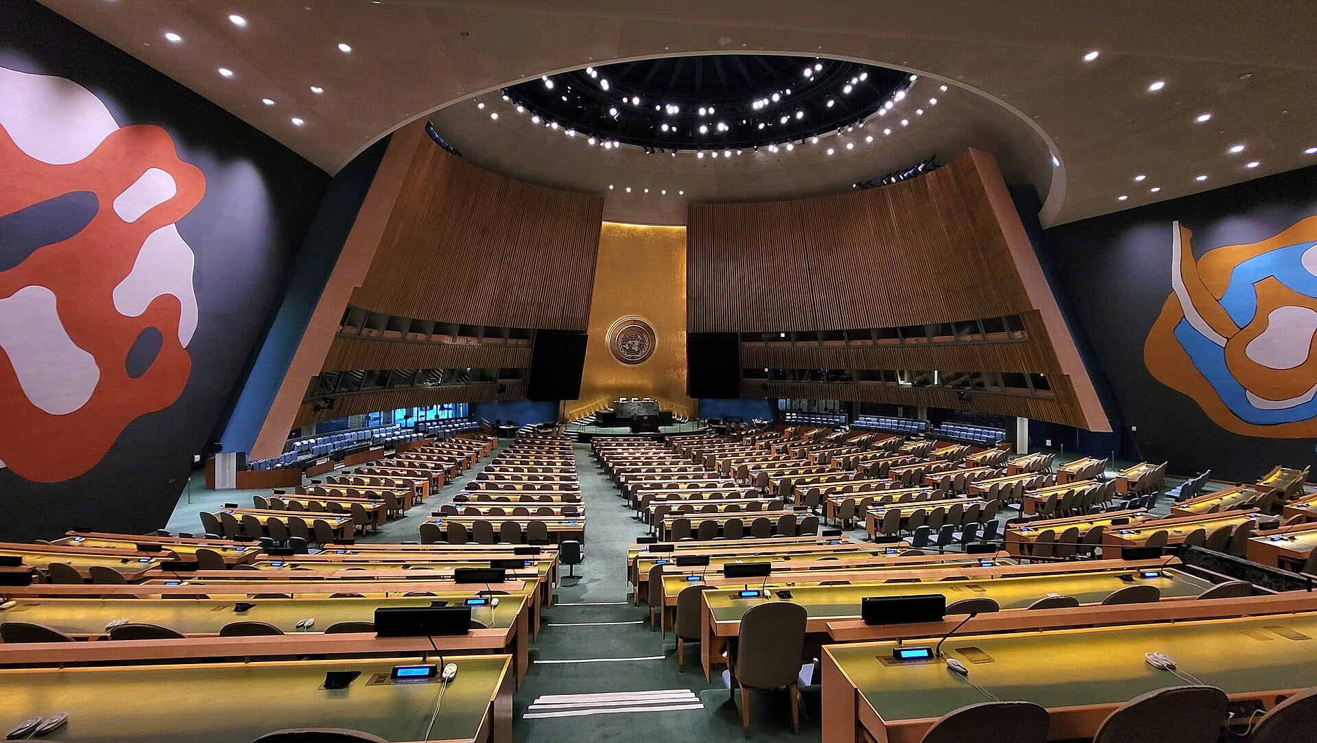 Empty United Nations General Assembly hall with the gold UN emblem on the podium wall and two large abstract murals on either side
