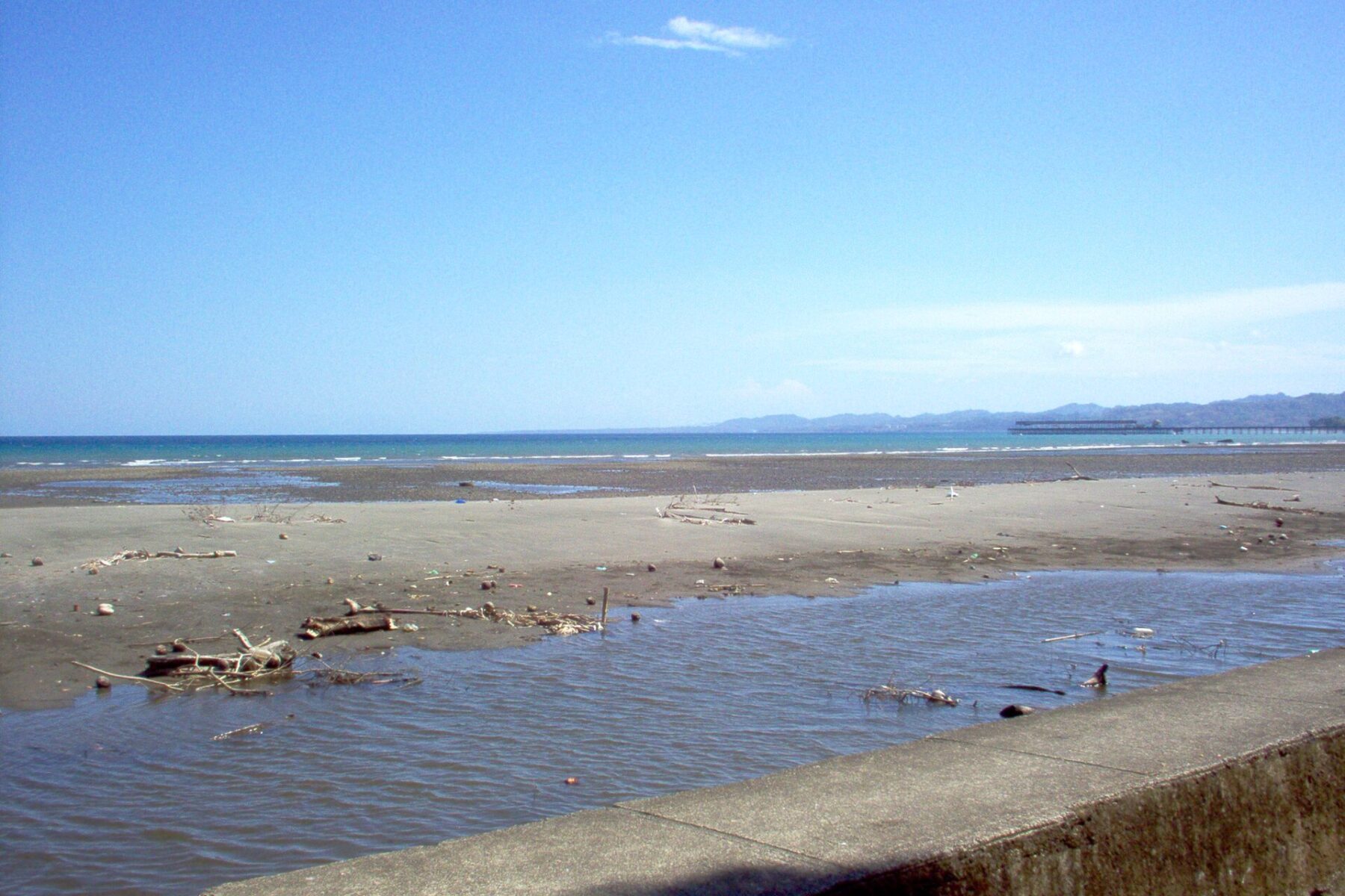 A grey sand beach at Puerto Armuelles in western Panama under a blue sky, with a port pier and the Punta Burica peninsula on the horizon
