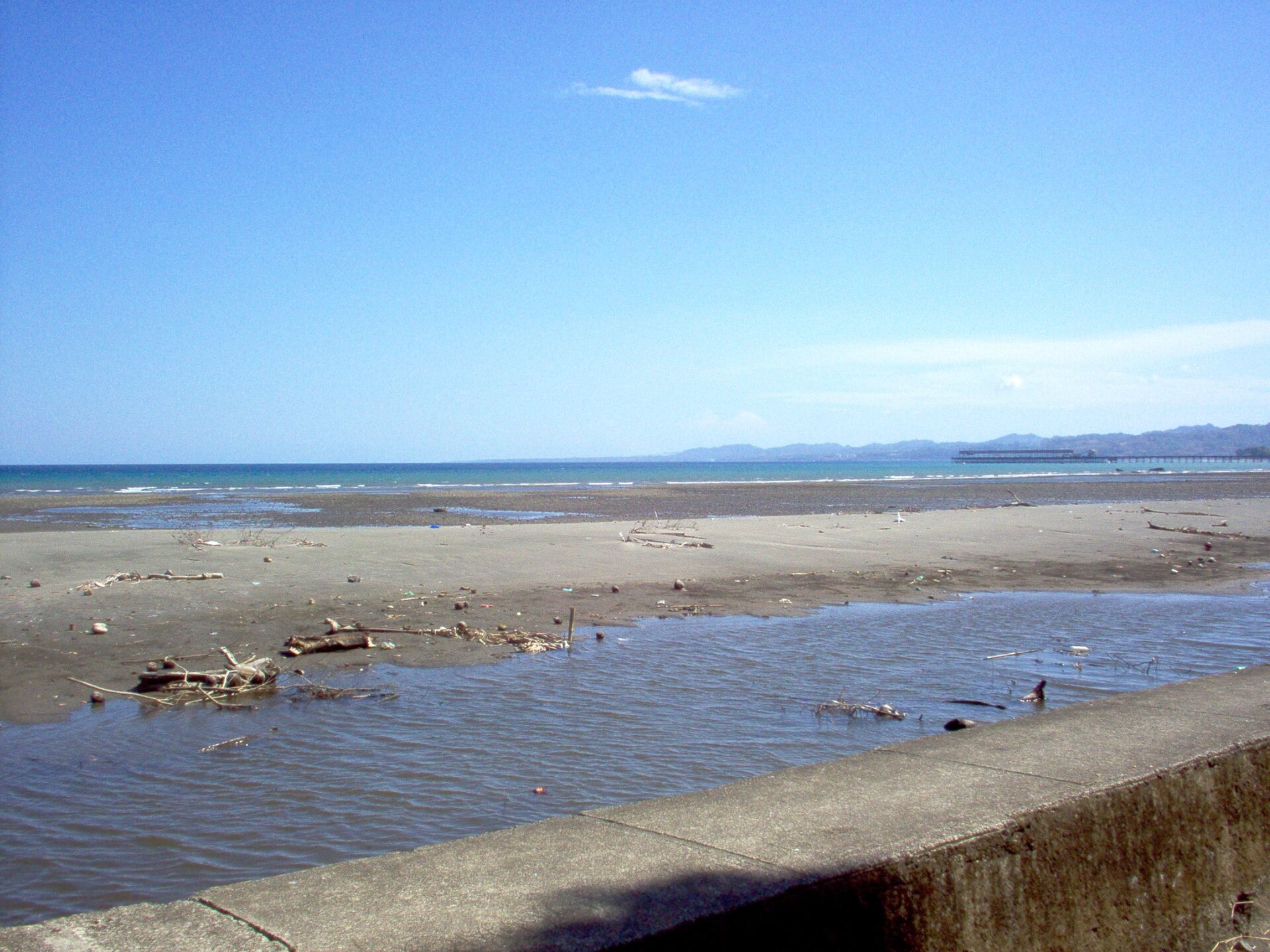 A grey sand beach at Puerto Armuelles in western Panama under a blue sky, with a port pier and the Punta Burica peninsula on the horizon