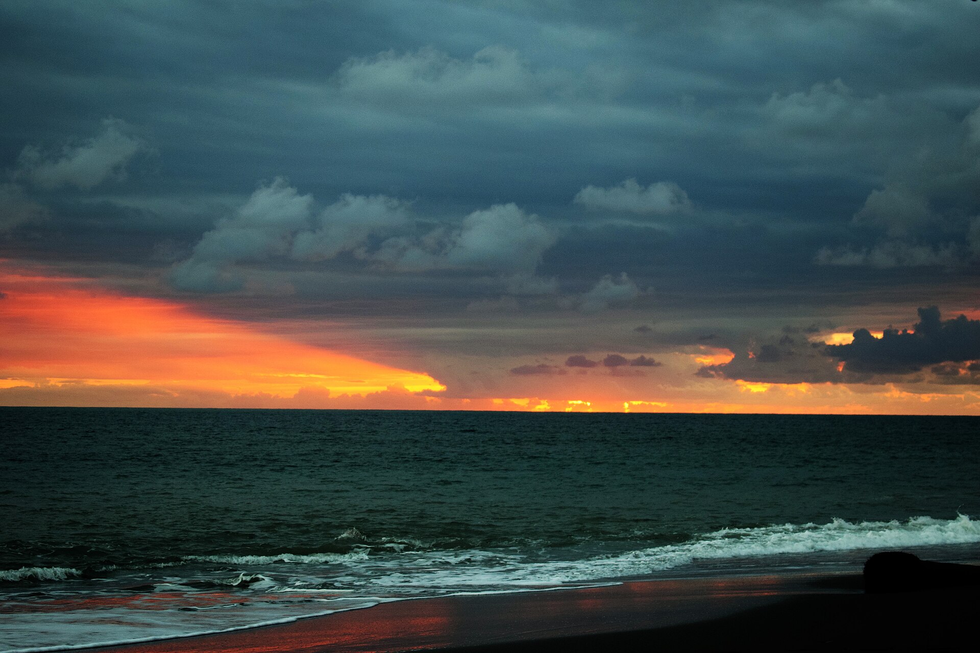An orange sunset under dark clouds over the Pacific Ocean, with waves breaking on a dark sand beach at Punta Burica