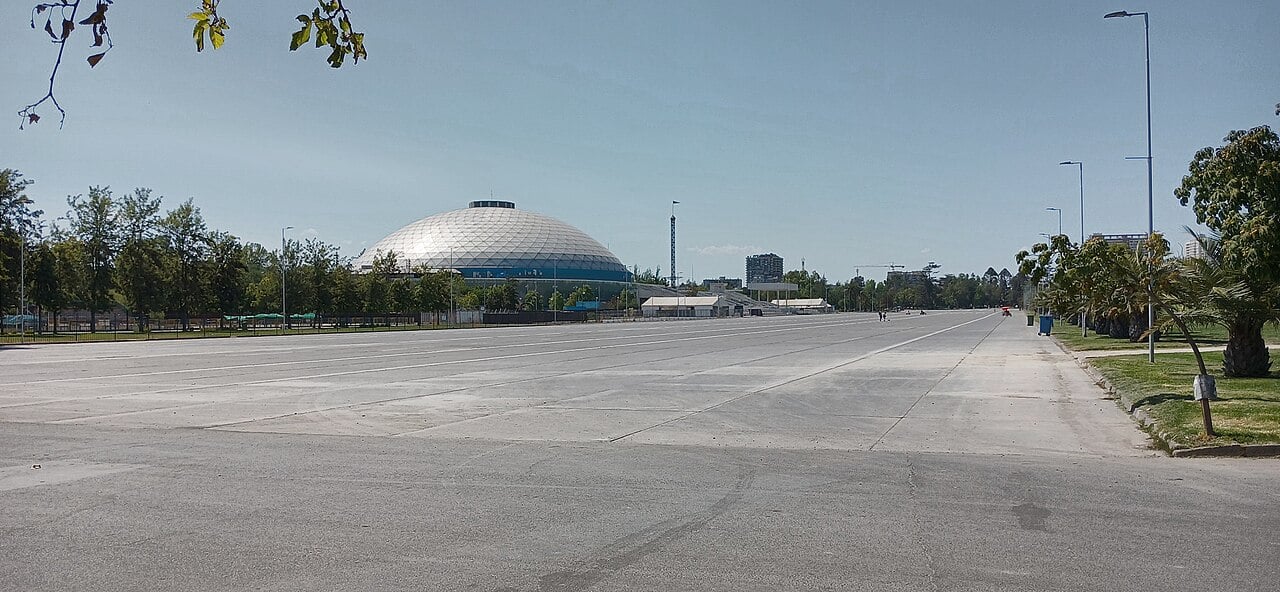 A white geodesic dome rises beyond a wide, empty paved esplanade lined with trees in Parque O'Higgins, Santiago, under a clear blue sky