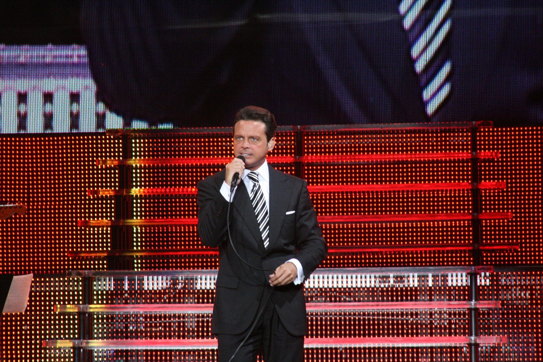 Luis Miguel singing on stage in a dark suit in front of a red lit backdrop
