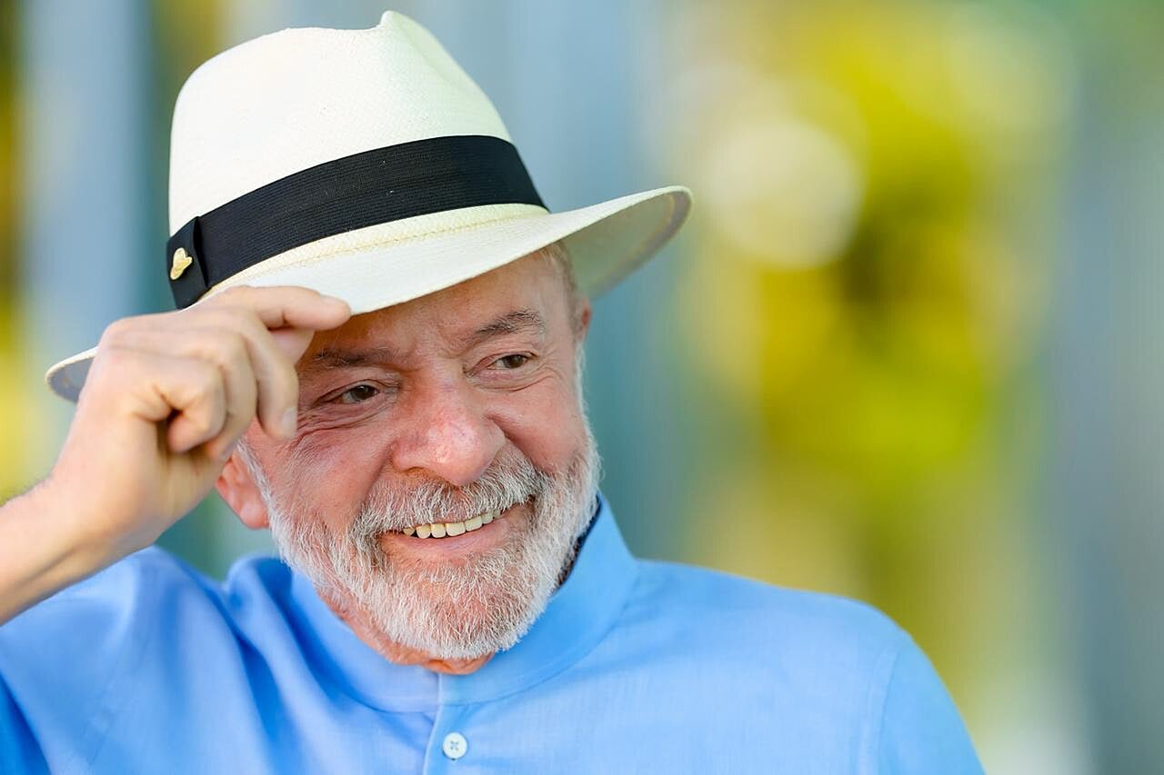President Lula in a white Panama hat and light blue shirt, smiling and touching the brim of his hat
