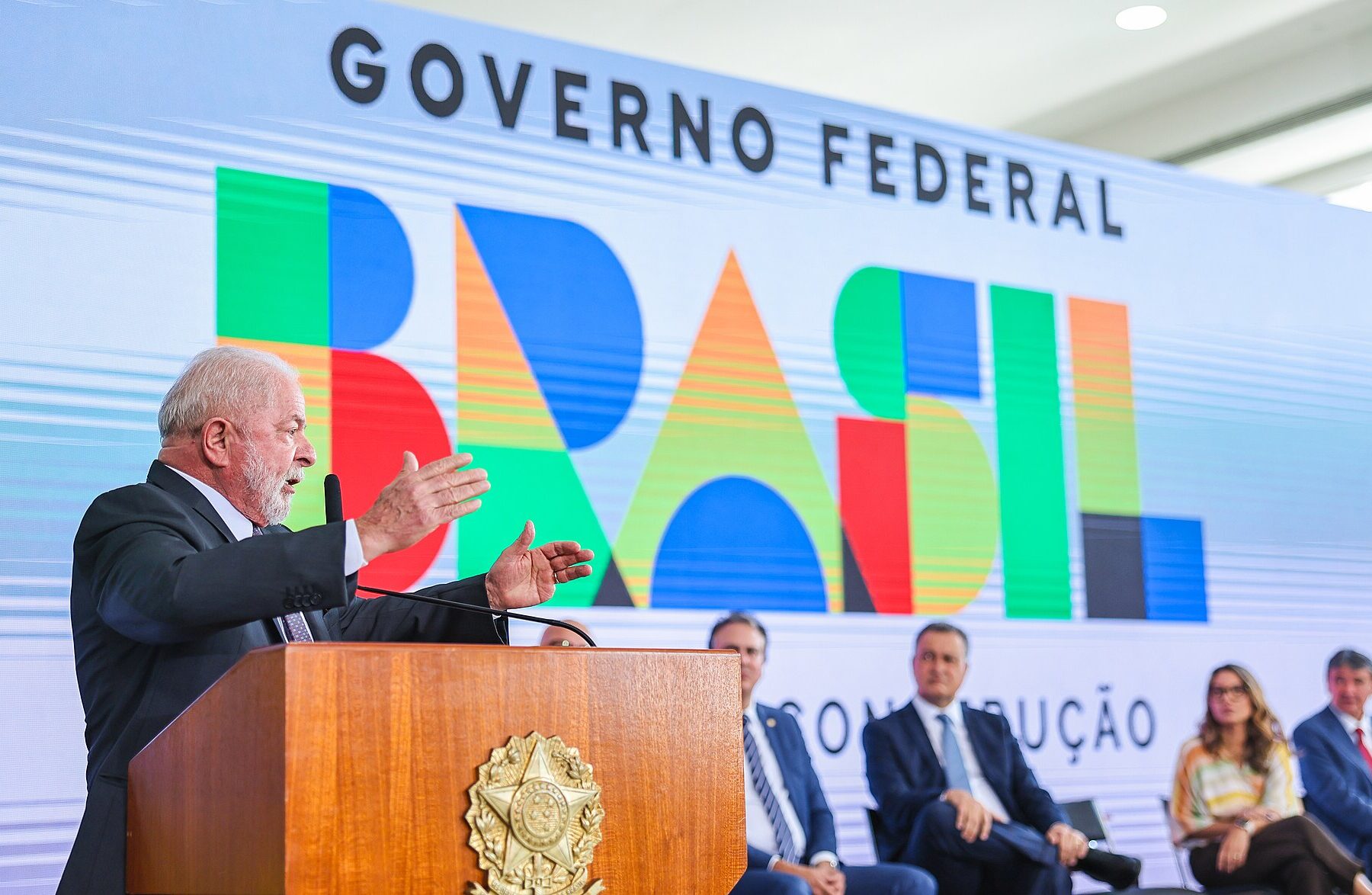 President Lula speaking at a wooden lectern with the presidential seal in front of a large Governo Federal Brasil banner, ministers seated beside him