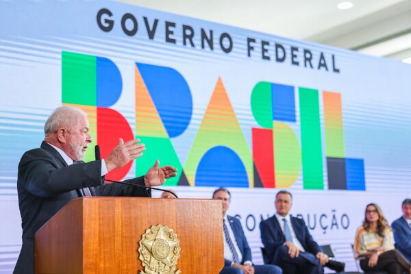 President Lula speaking at a wooden lectern with the presidential seal in front of a large Governo Federal Brasil banner, ministers seated beside him