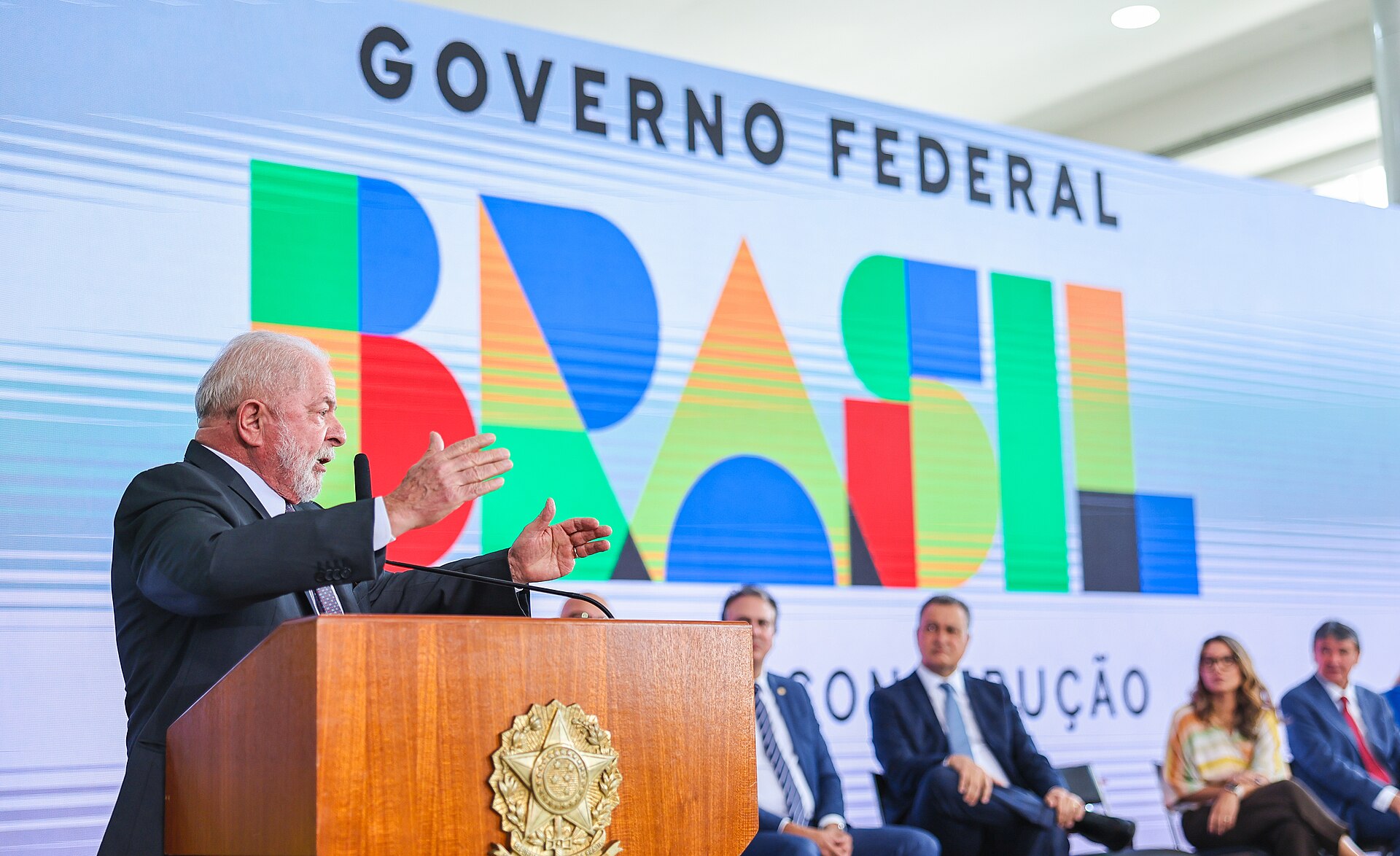President Lula speaking at a wooden lectern with the presidential seal in front of a large Governo Federal Brasil banner, ministers seated beside him