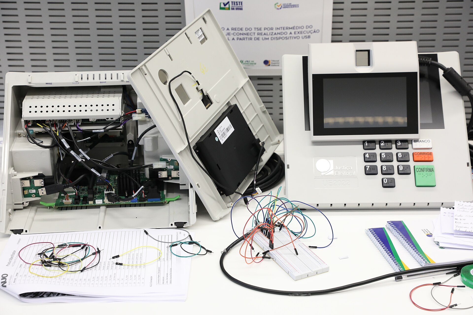 A Brazilian electronic voting machine with its casing open beside a second machine with a screen and keypad on a test table
