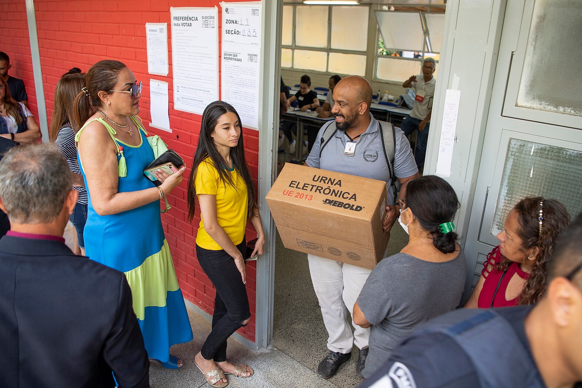An election official carries a cardboard box marked electronic voting machine through a doorway as voters wait beside a red brick wall with voting notices