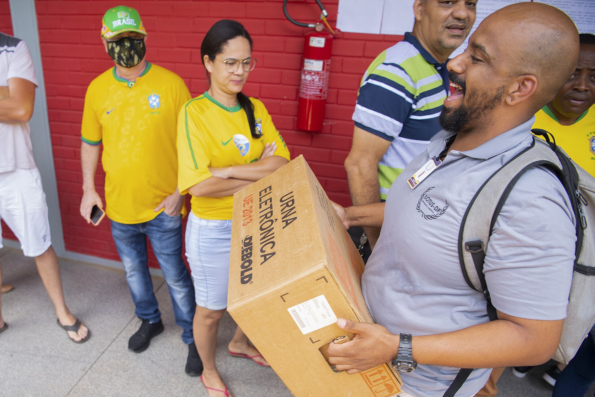 A smiling election official carries a cardboard box marked electronic voting machine while voters in yellow Brazil shirts wait beside a red brick wall