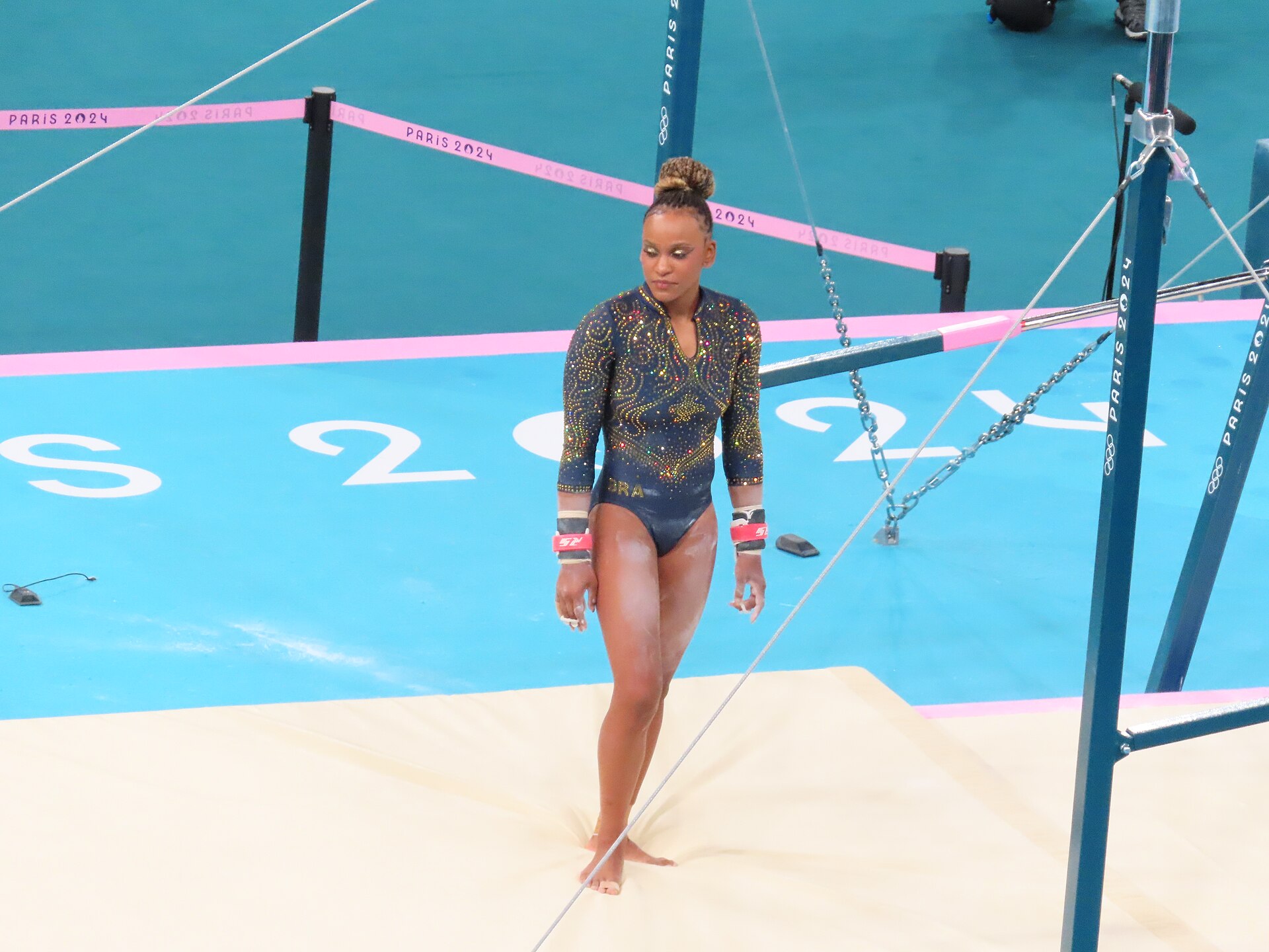 Gymnast in a dark sparkling leotard walks across a mat beside the uneven bars at the Paris 2024 Olympics
