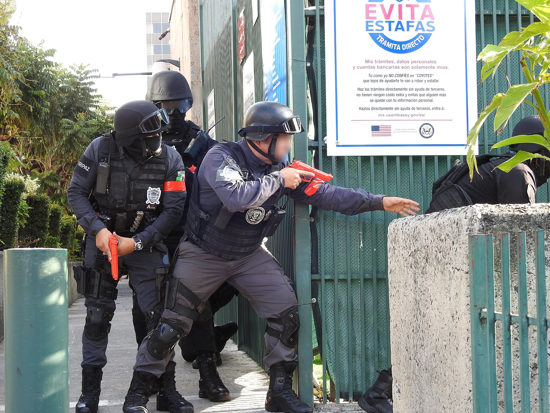 Masked Guadalajara municipal police SWAT officers in helmets and body armour aim orange training pistols along a green fence at a US consulate training exercise