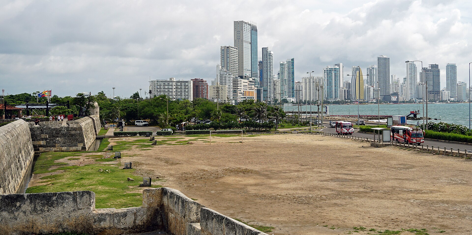 Tall residential towers of Cartagena's Bocagrande district across the bay, seen over an old stone bastion and open dry ground under a cloudy sky