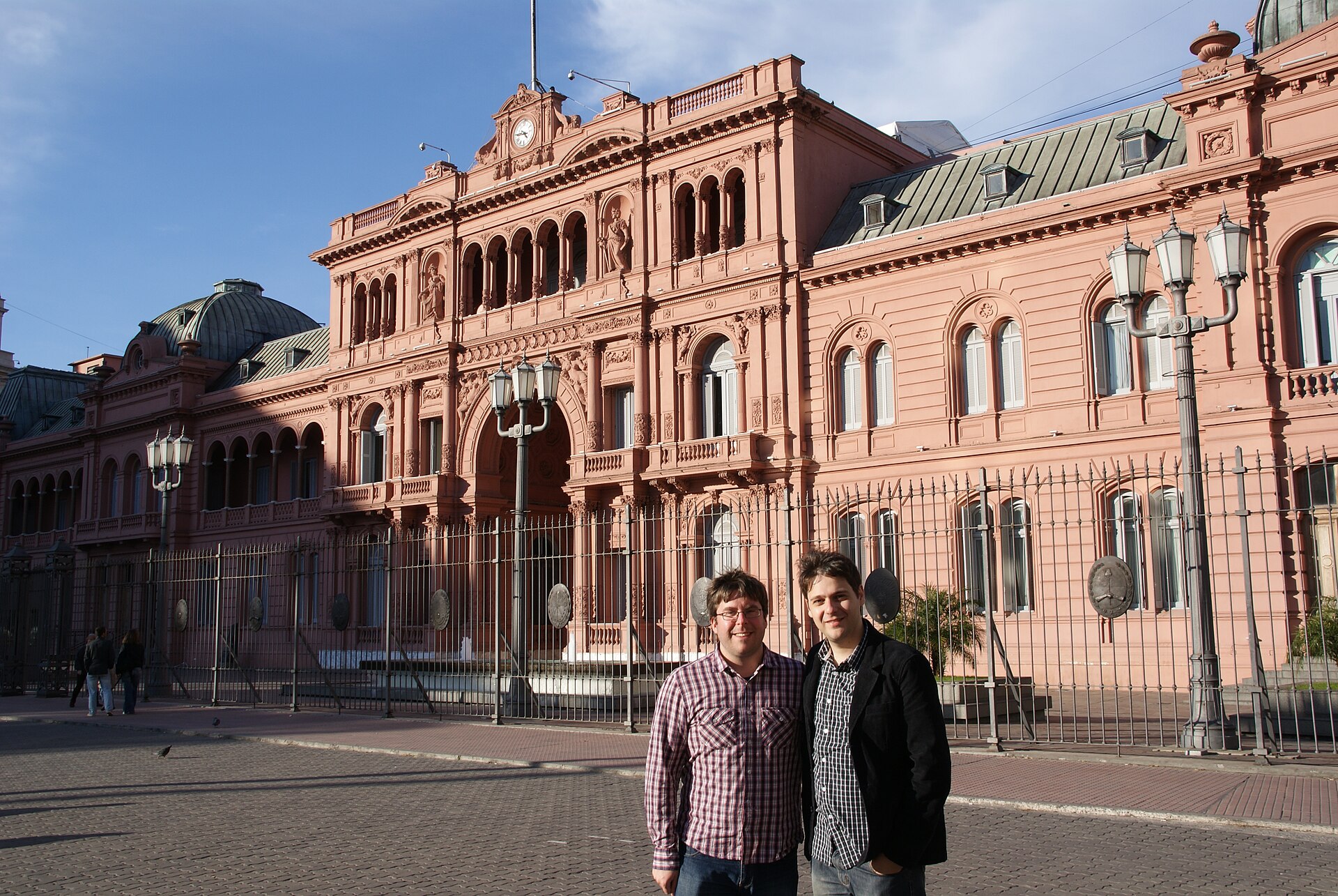 The pink facade of the Casa Rosada behind an iron fence, with two men posing in the foreground
