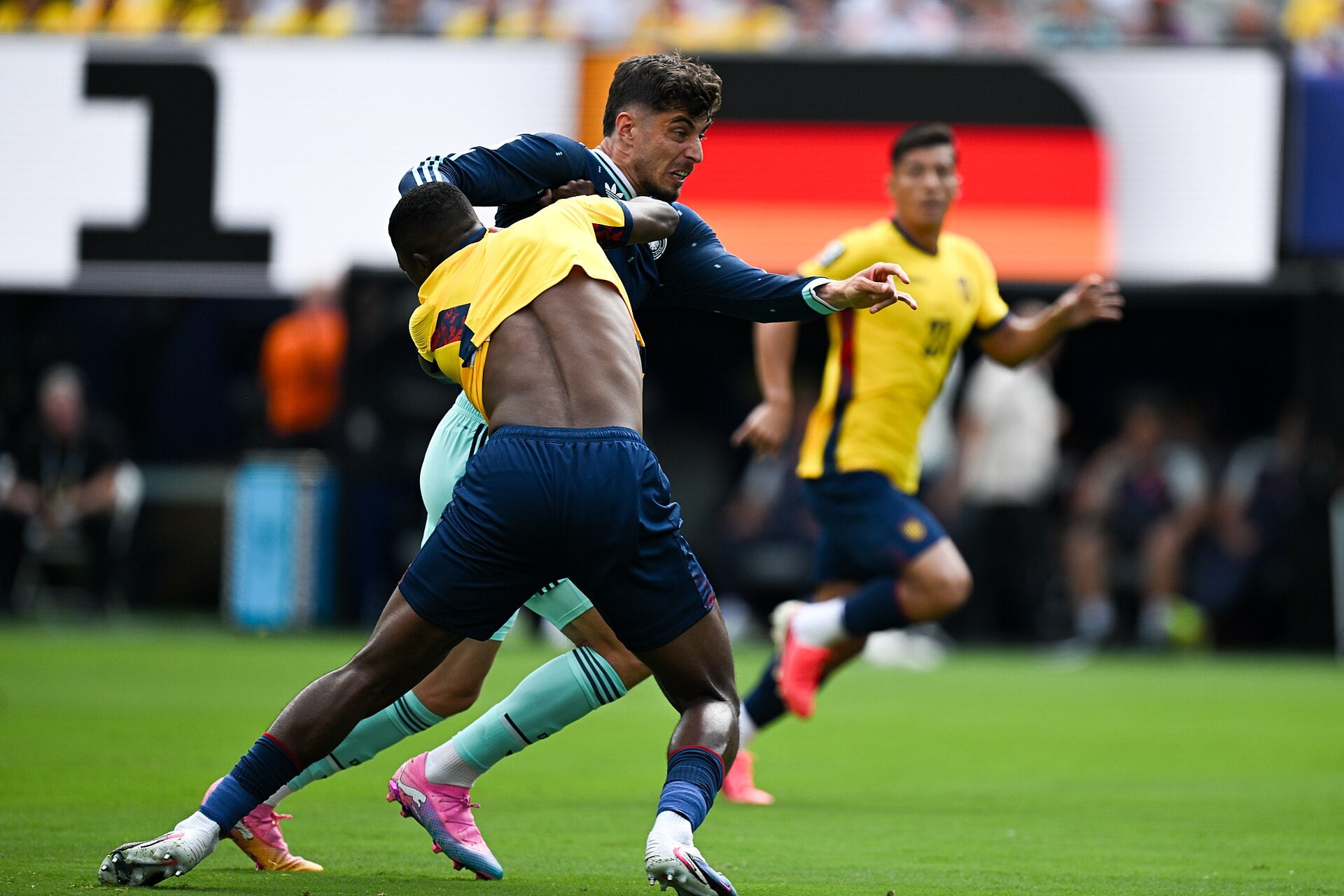 Ecuador defender Willian Pacho in a yellow shirt and Germany's Kai Havertz in a dark shirt challenge for the ball on a grass pitch during a World Cup match