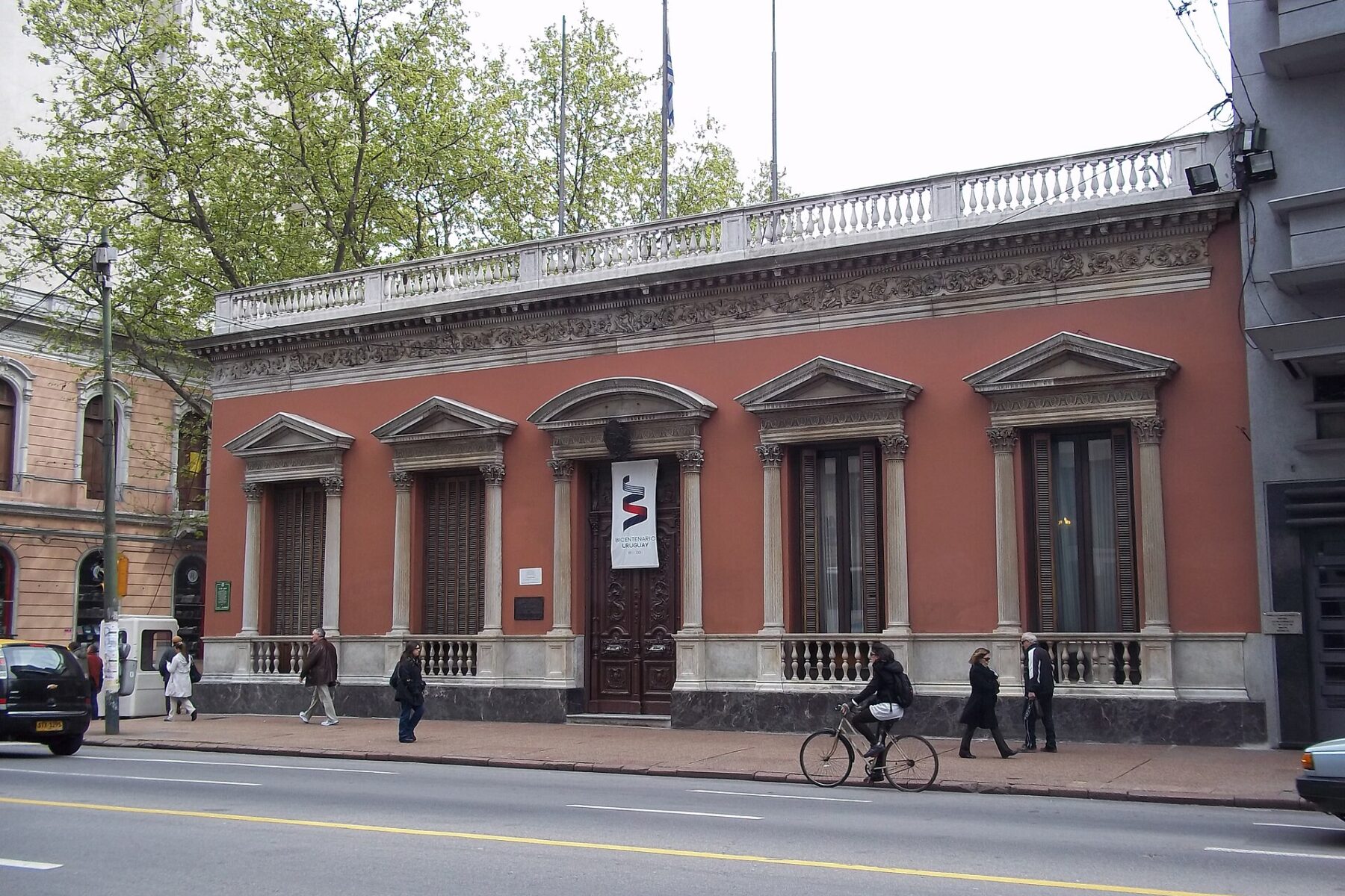 Pink two-storey neoclassical building with tall windows and columns on a Montevideo avenue, with pedestrians and a cyclist on the pavement