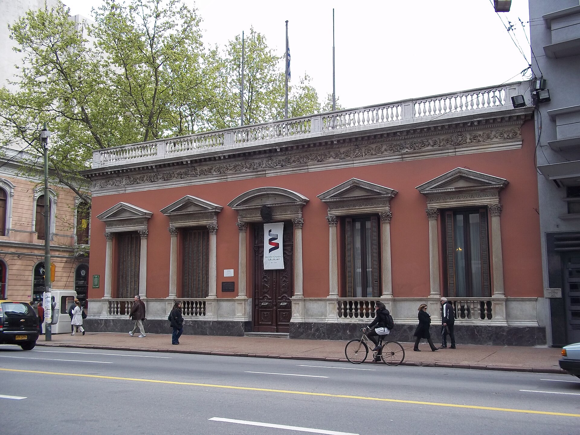 Pink two-storey neoclassical building with tall windows and columns on a Montevideo avenue, with pedestrians and a cyclist on the pavement