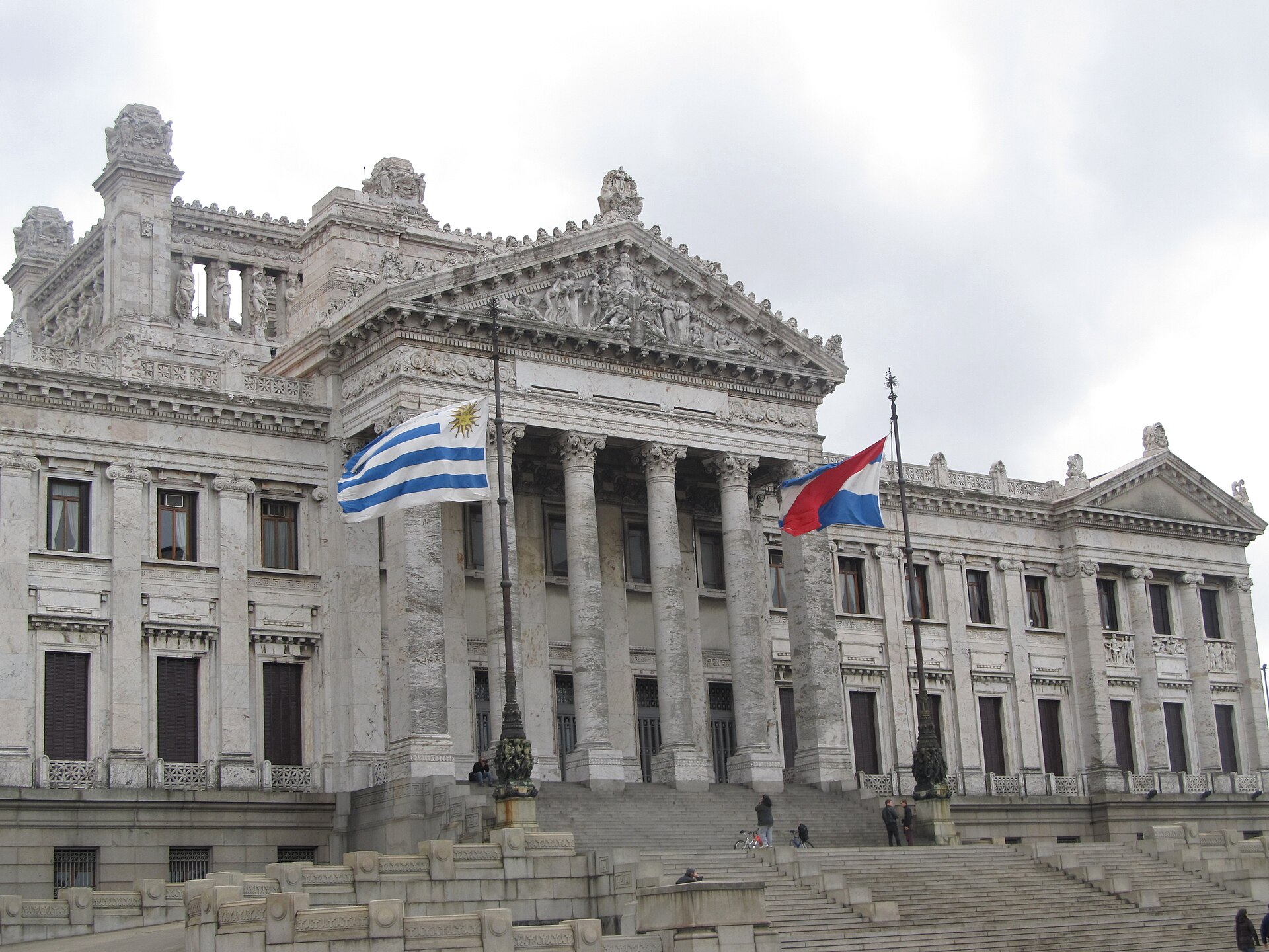 White marble Legislative Palace in Montevideo with Uruguayan and Artigas flags flying in front of its columned portico under a cloudy sky