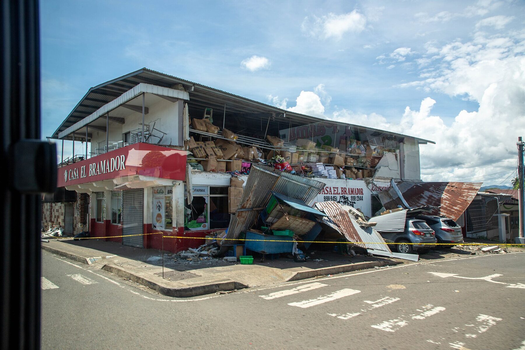 A two-storey corner shop named Casa El Bramador with its upper floor torn open and its metal awning fallen onto the pavement behind yellow tape in Soná, Panama