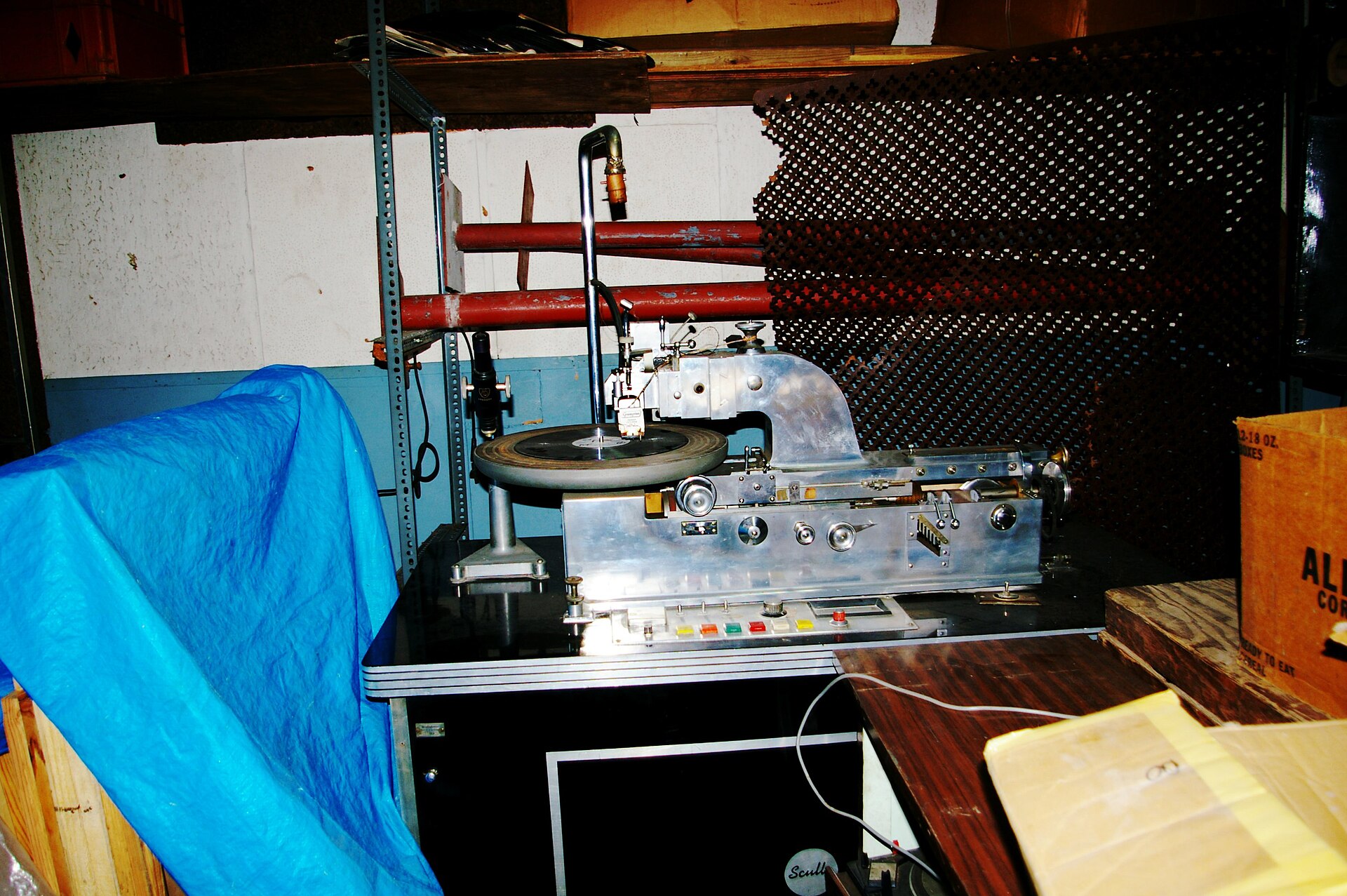 A silver vinyl record cutting lathe with a disc on the turntable, in a cluttered basement room with a blue tarpaulin beside it
