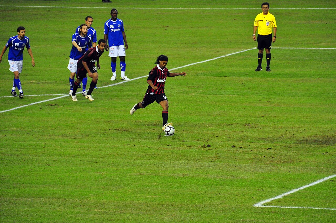 Footballer in a red and black AC Milan shirt strikes a penalty on a grass pitch while players and a referee in yellow look on