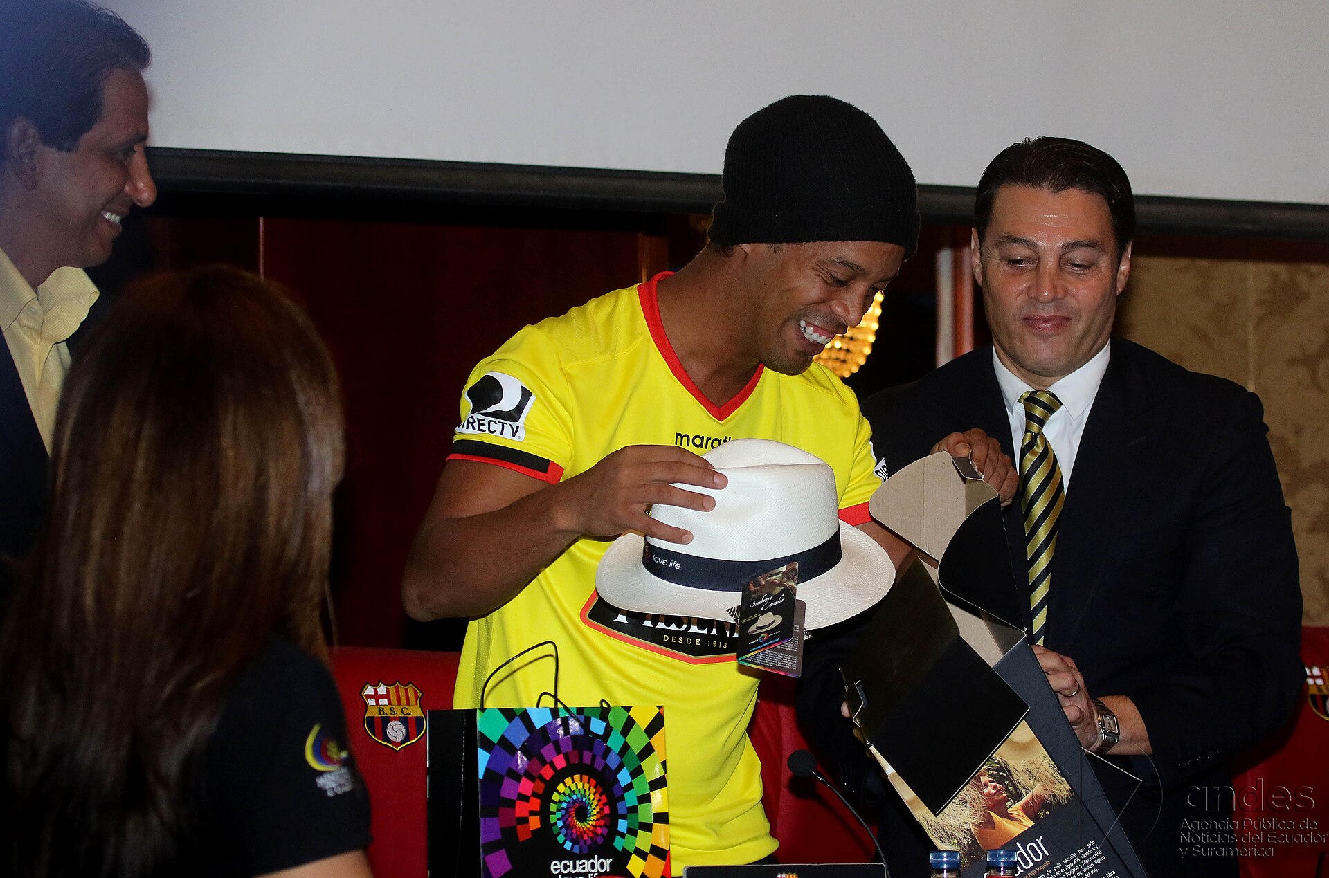 Smiling man in a yellow football shirt and black beanie holds a white straw hat at a press event in Guayaquil
