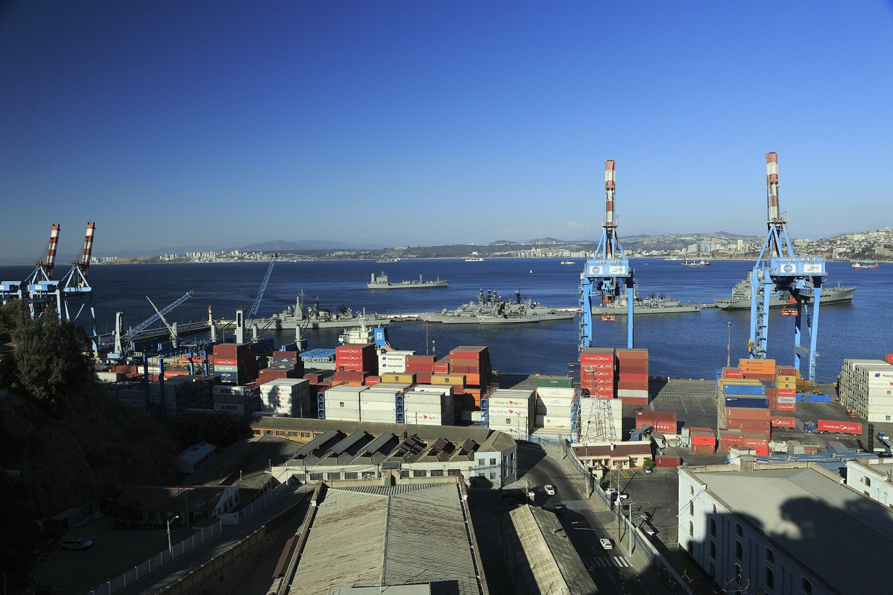 Container stacks and two blue gantry cranes at the port of Valparaíso, with grey naval ships moored in the bay behind