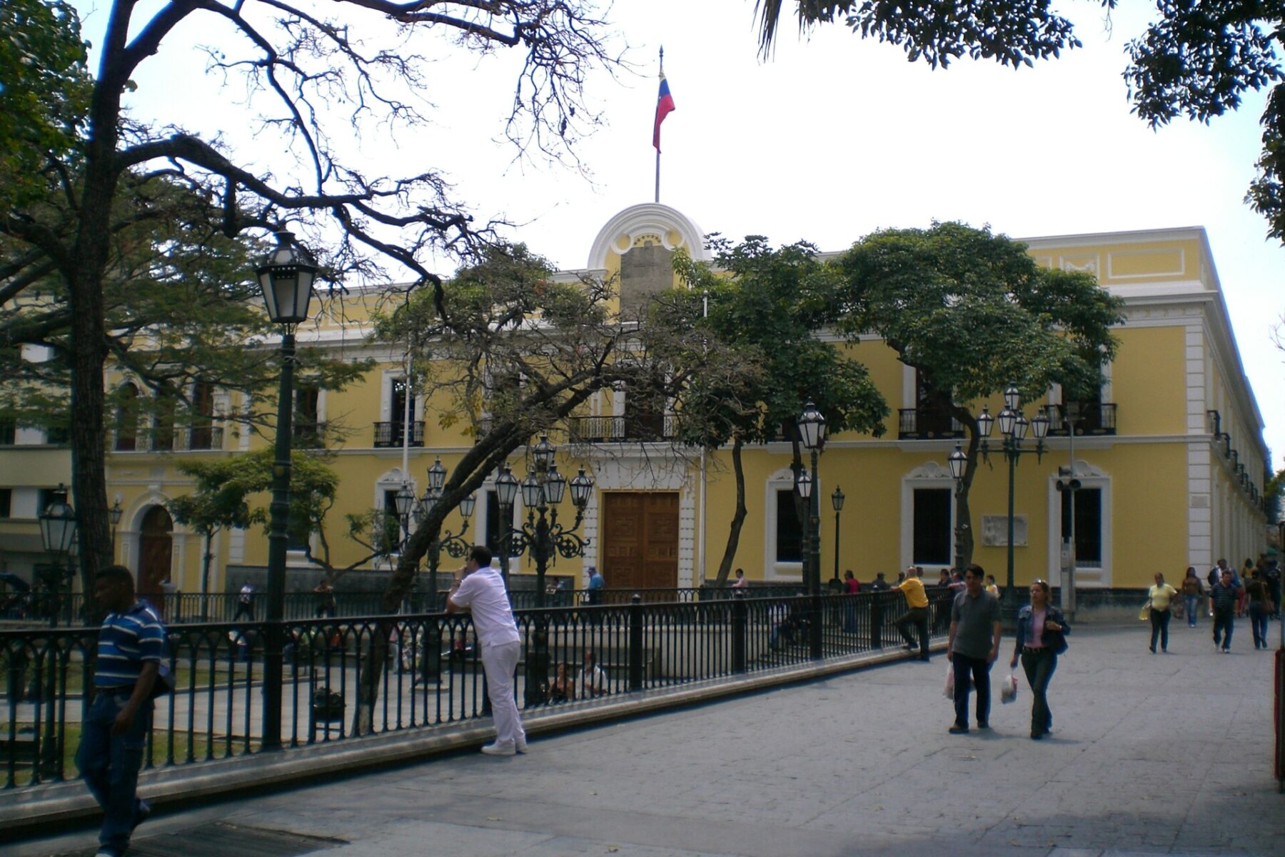 The yellow colonial facade of the Casa Amarilla in Caracas, with a Venezuelan flag on the roof, trees and people walking on the square in front