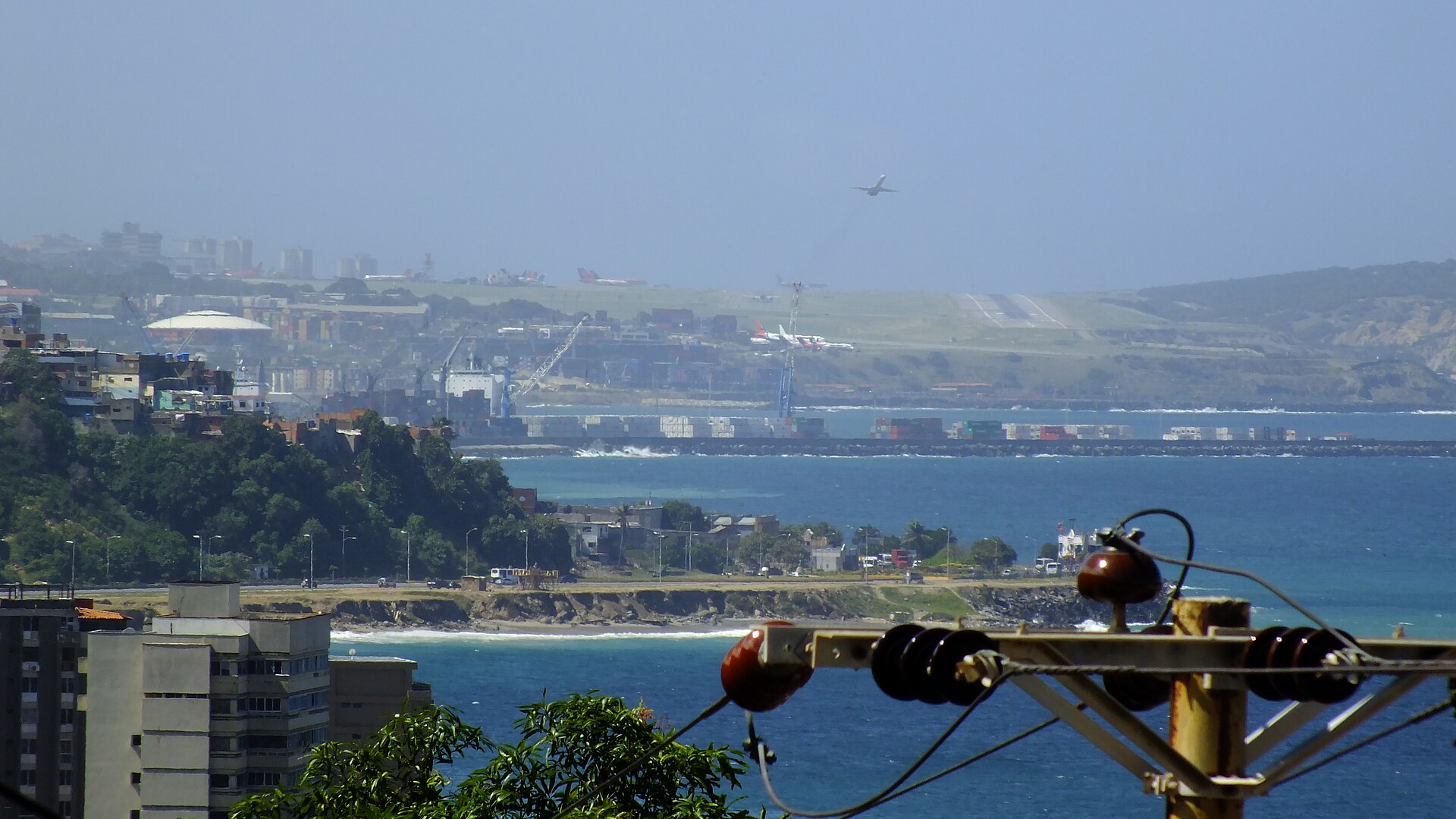 View across the sea from Macuto to the container port of La Guaira, with cranes and stacked containers, and Maiquetía airport behind it as a plane takes off