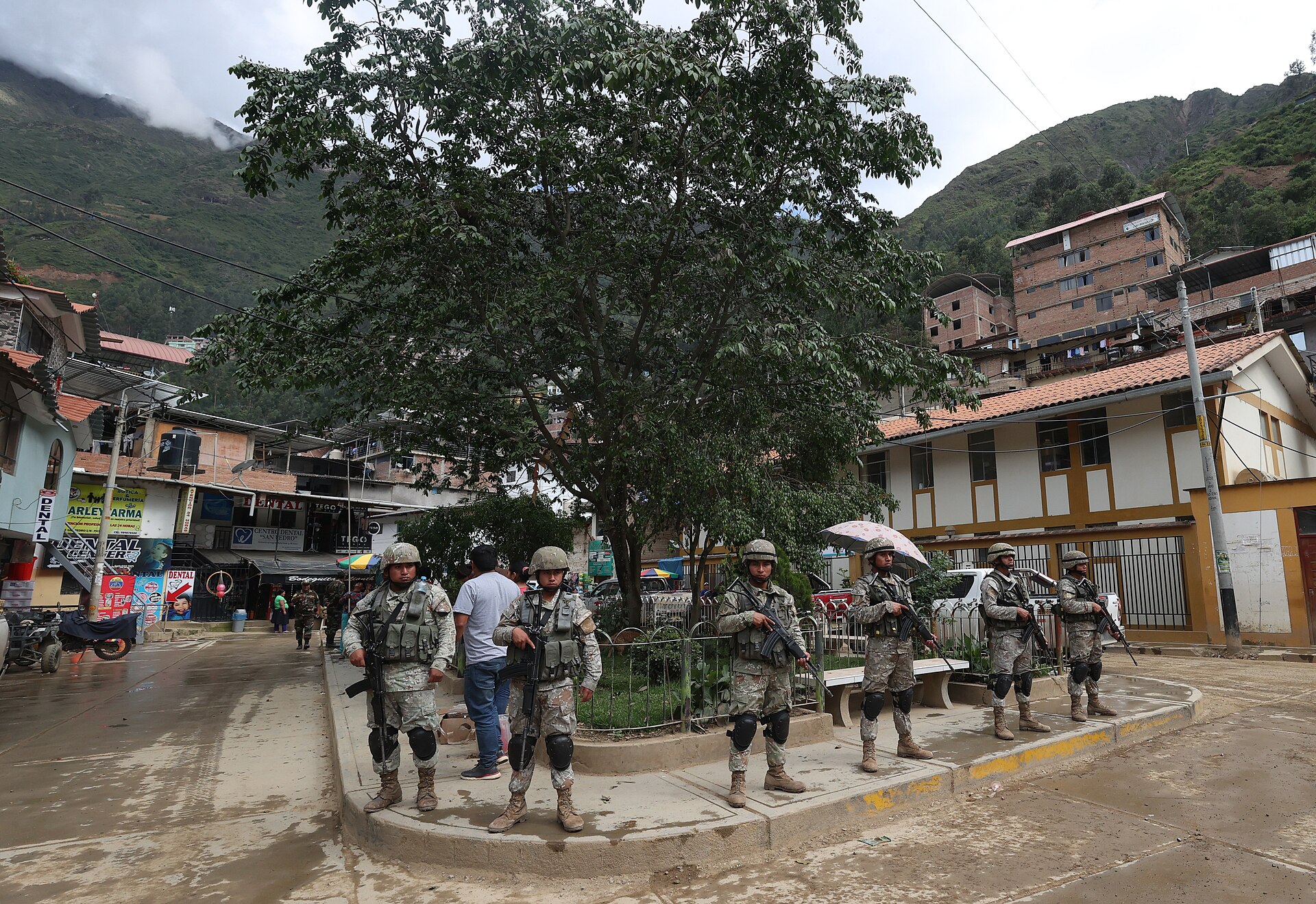 Armed soldiers stand around a small tree-shaded square in a mountain town, with green hills behind