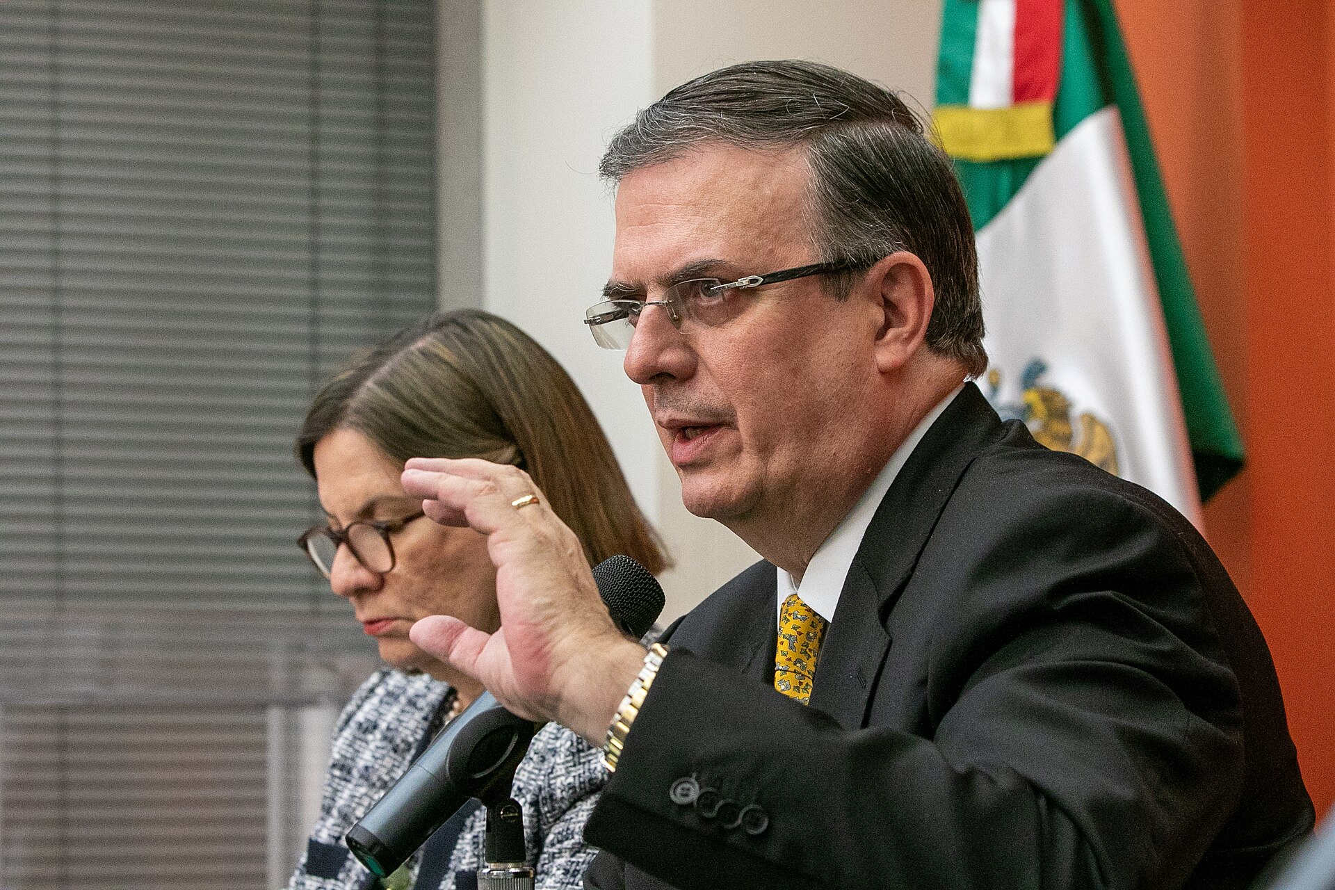 Marcelo Ebrard speaking into a microphone in front of a Mexican flag, with a woman seated beside him