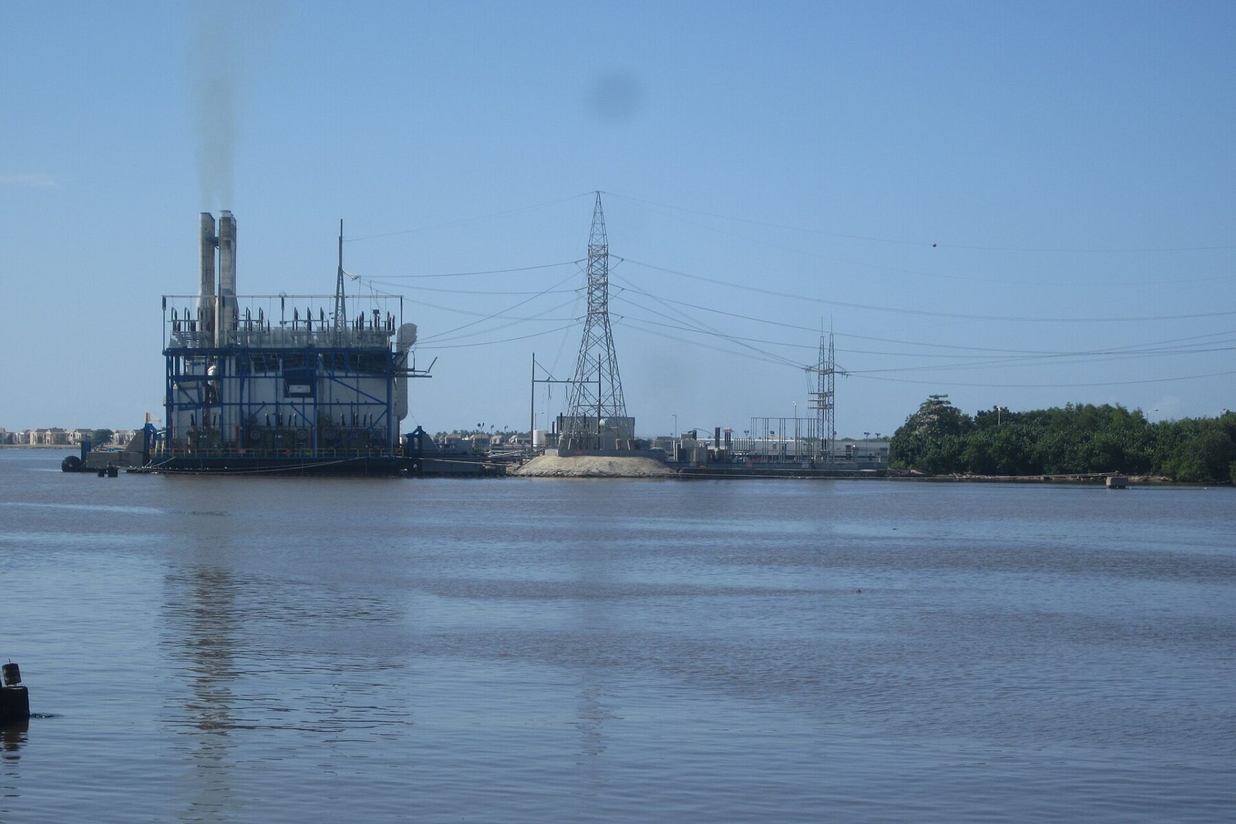 A floating power plant with two smokestacks sits on the Higuamo River beside a steel transmission tower and power lines, Dominican Republic.