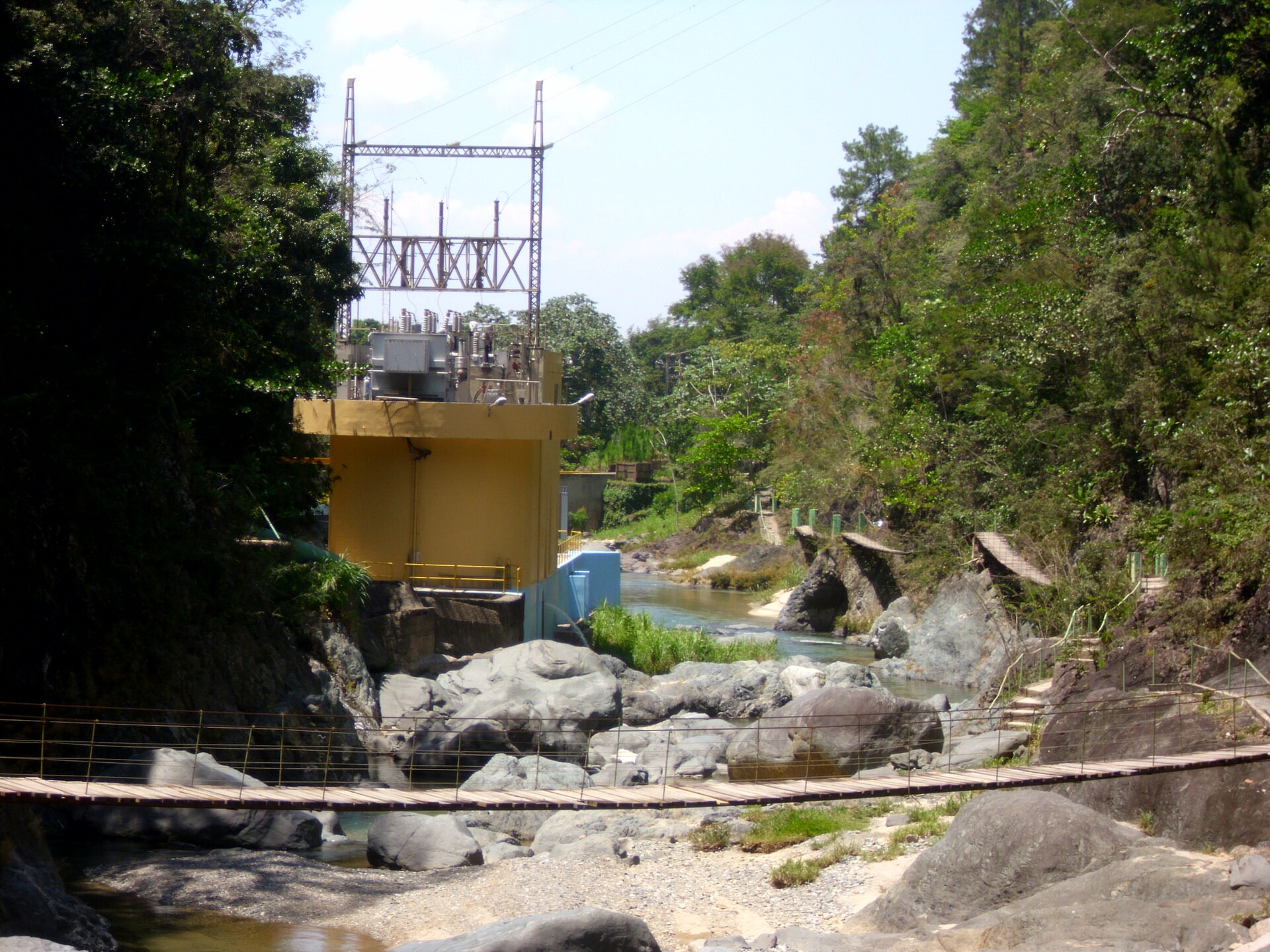 A yellow hydroelectric station building with a transformer yard on its roof stands beside a rocky river, with a footbridge in front.