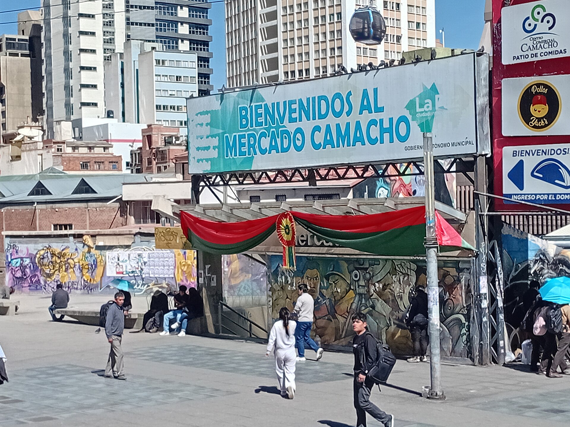 Pedestrians walk past the entrance sign of Mercado Camacho, with murals and office towers behind, in La Paz