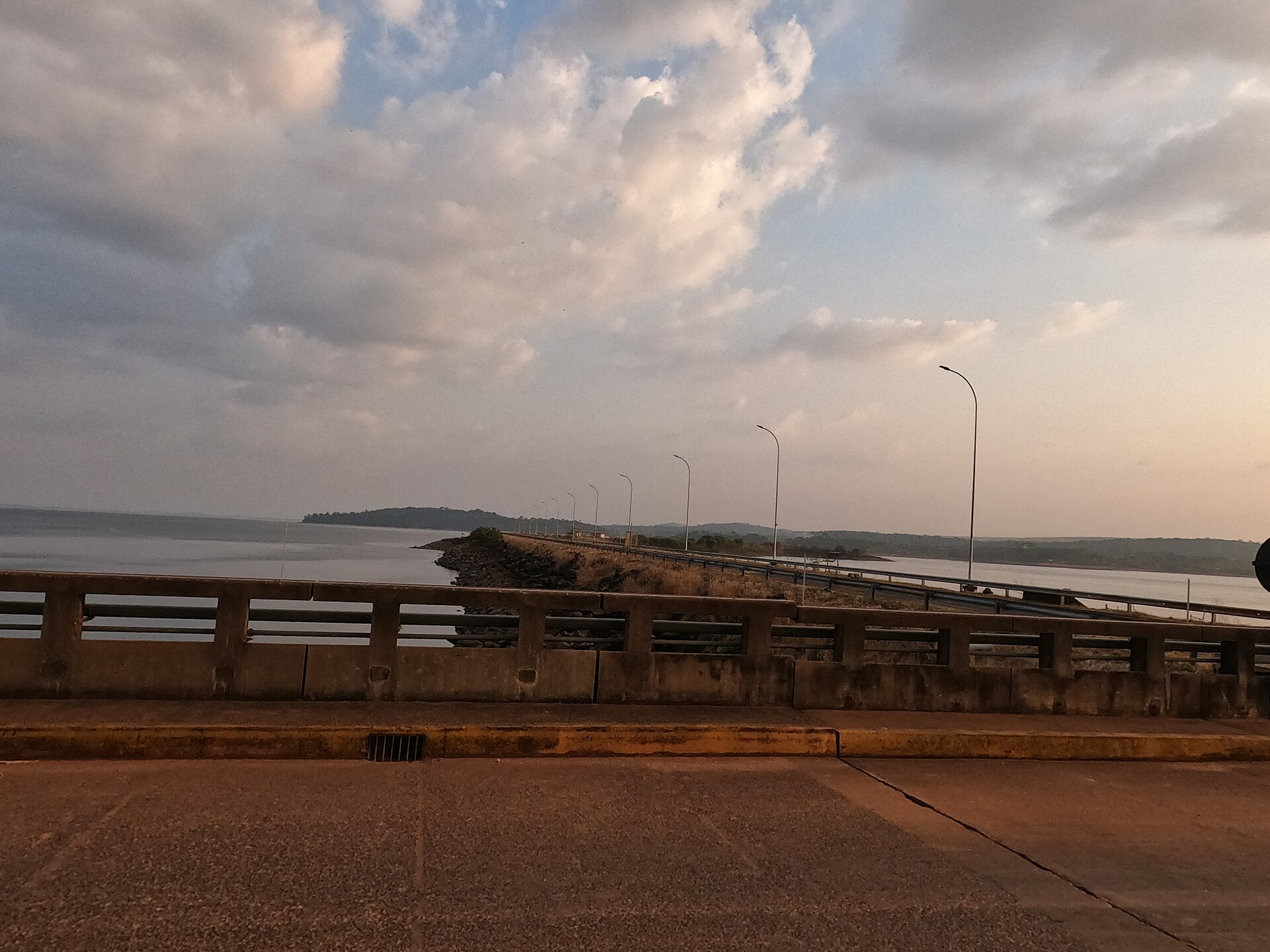 A road with street lamps runs along the crest of a long dam beside a wide reservoir under a cloudy evening sky