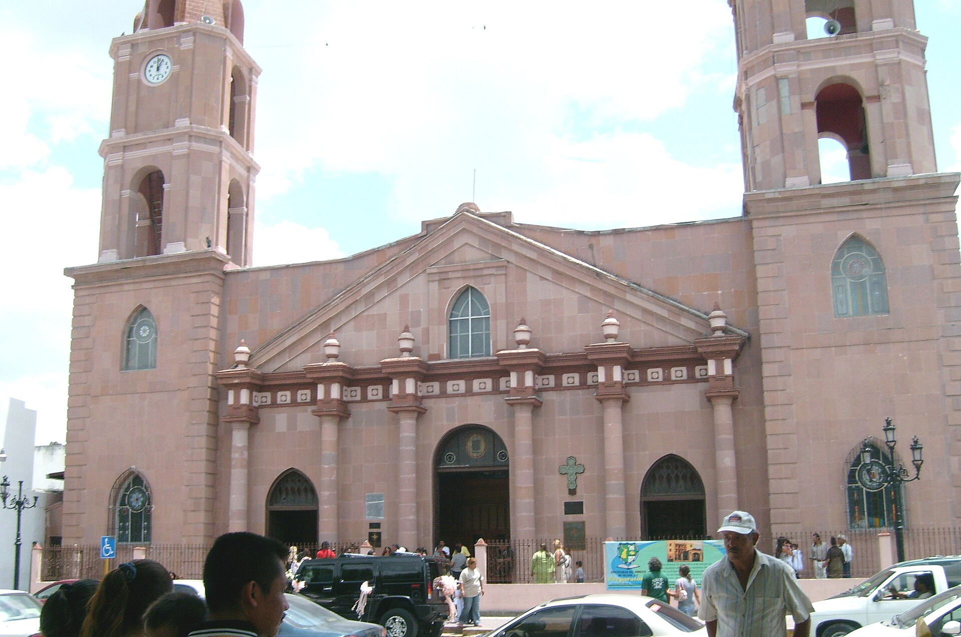 Pink stone facade and twin bell towers of the Cathedral of Our Lady of Refuge in Matamoros, with people and cars in front