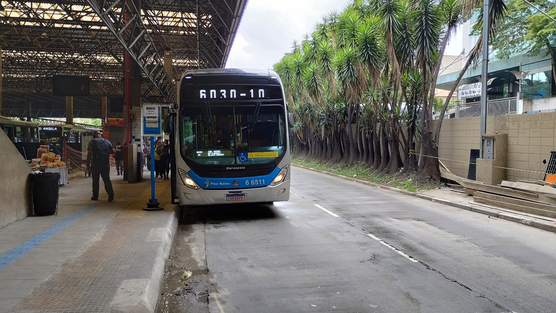 A white and blue battery-electric city bus showing route 6030-10 stops beside a covered bus terminal platform in São Paulo