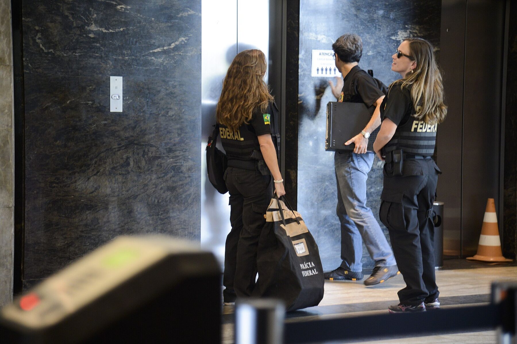 Brazilian Federal Police agents, two in black vests marked Federal, wait at a lift in an office lobby with an evidence bag