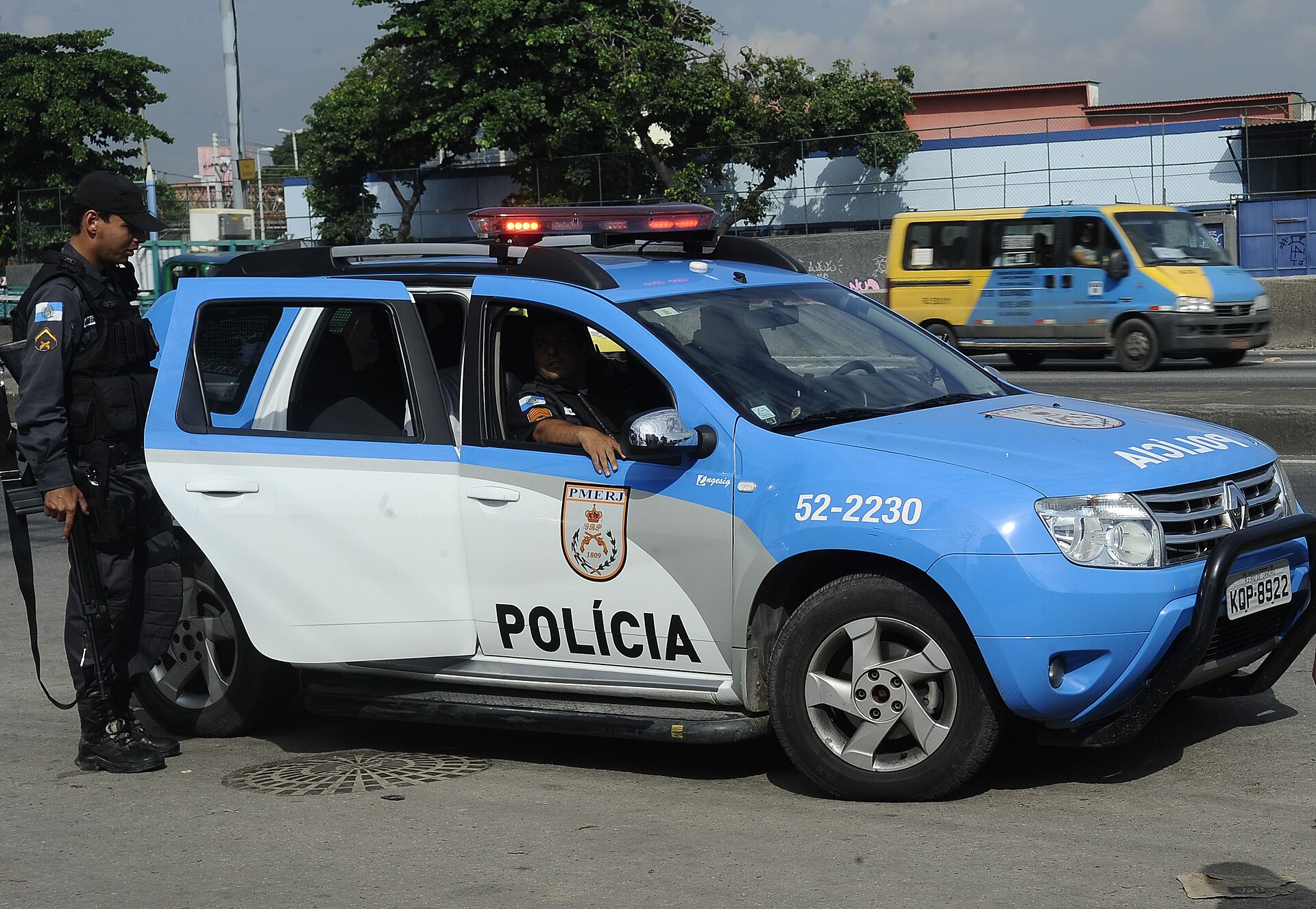 A Rio de Janeiro military police officer in a black uniform stands beside a blue and white patrol car on a city road