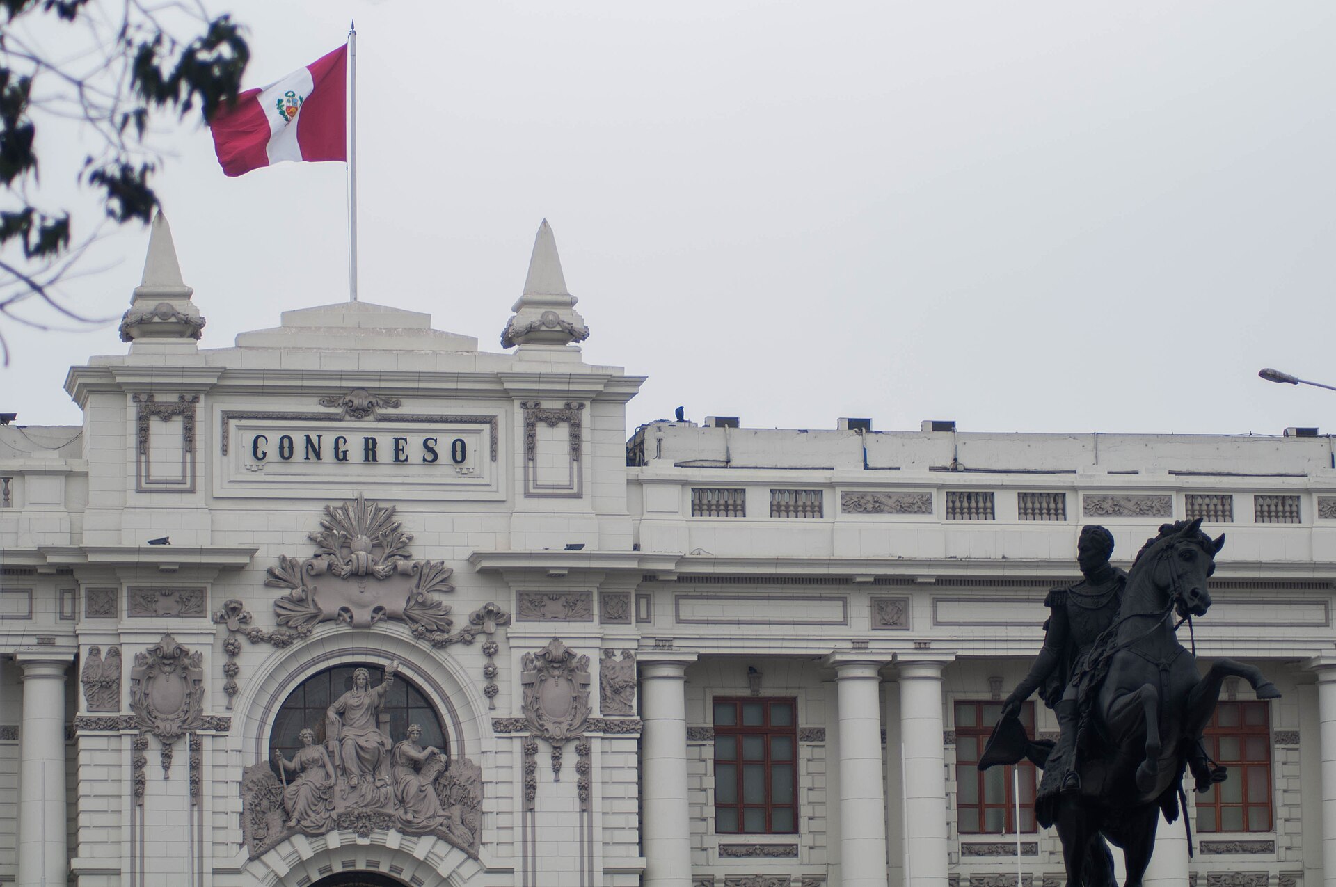 White neoclassical facade of Peru's Congress building with the word Congreso, a Peruvian flag on the roof and a dark equestrian statue in the foreground