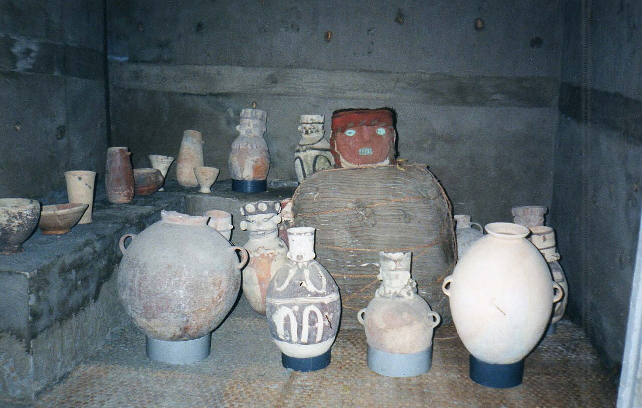 Pre-Hispanic ceramic jars and vessels arranged on a mat around a wrapped funerary bundle with a painted red mask, in a concrete display room