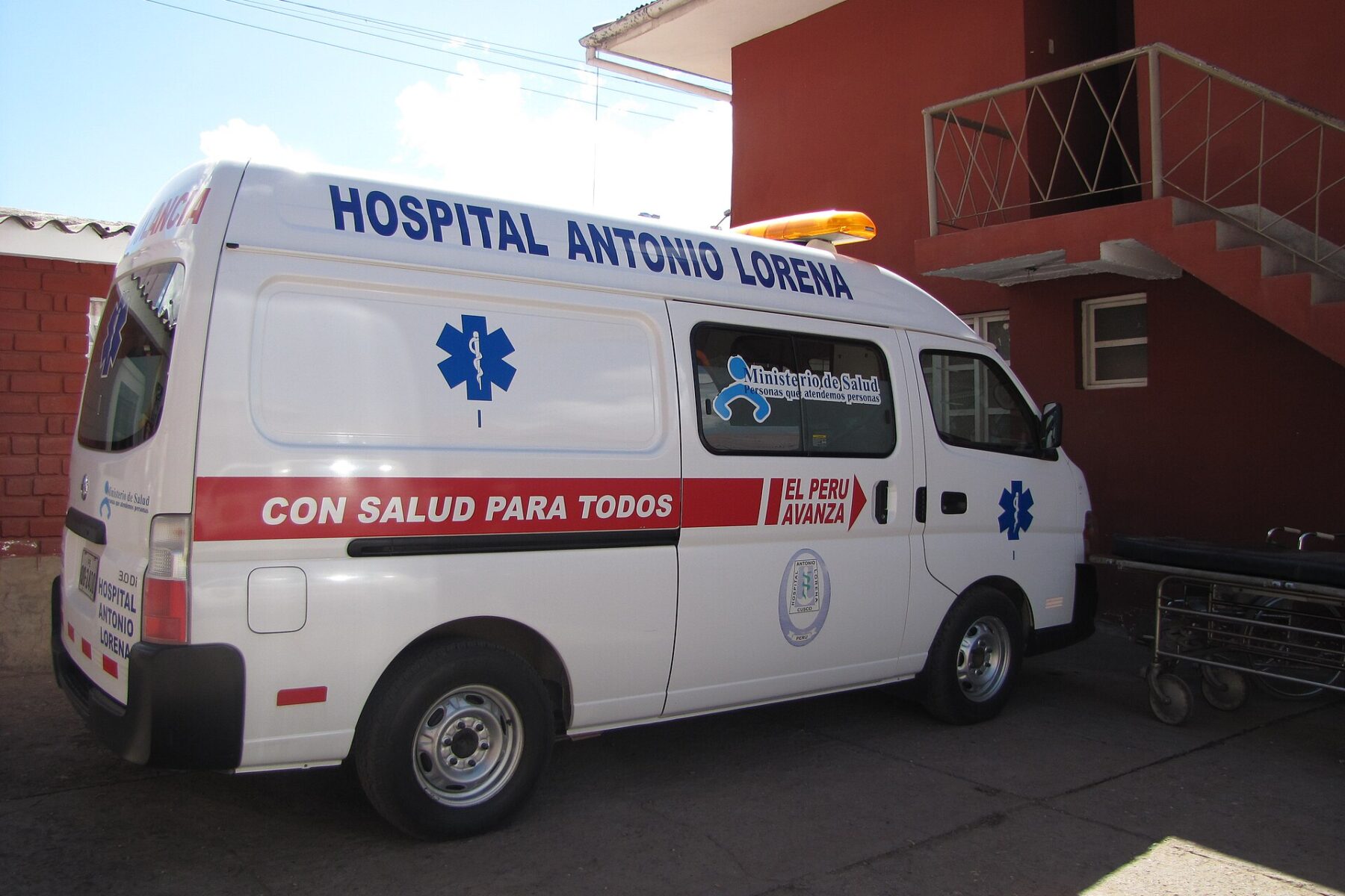A white Peruvian health ministry ambulance marked Hospital Antonio Lorena parked outside a red-painted building with an outdoor staircase in Cusco