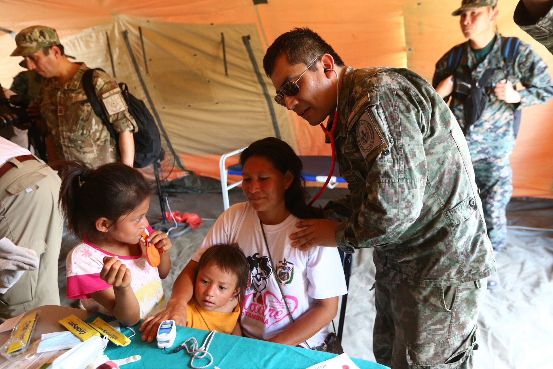 A Peruvian army medic in camouflage with a stethoscope examines a woman holding a small child, beside a girl, at a table inside an orange field tent
